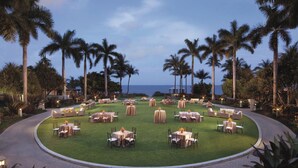 A circular outdoor dining area with tables and chairs, surrounded by palm trees and a view of the ocean.