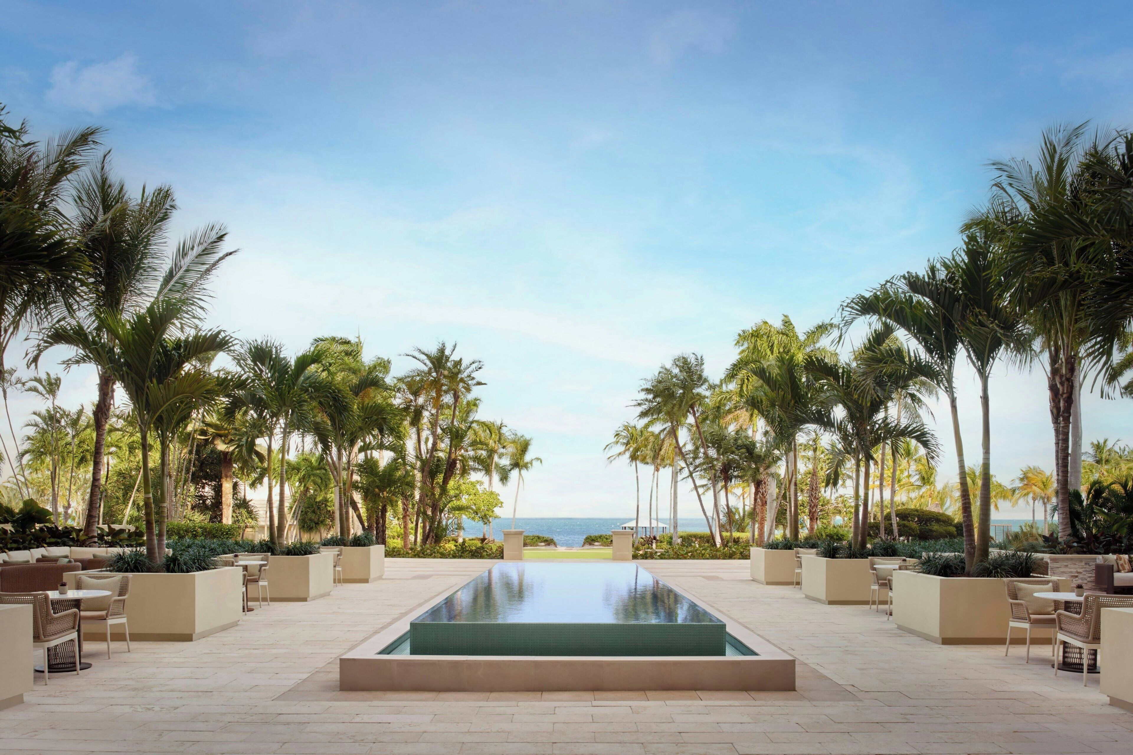 A pool area with a view of the ocean, surrounded by palm trees and outdoor seating.