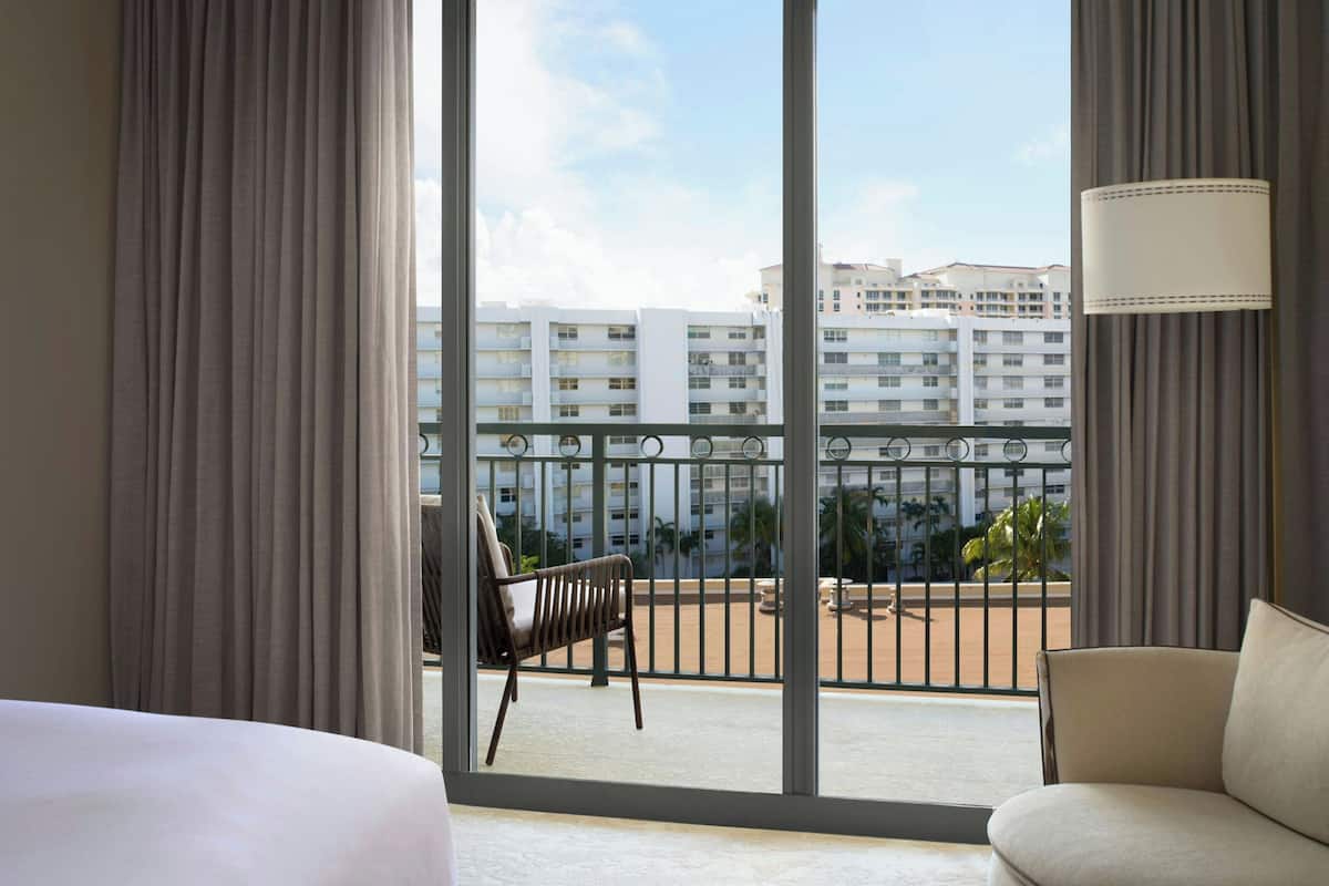 A hotel room with a large window, a balcony with a chair, and a view of high-rise buildings.