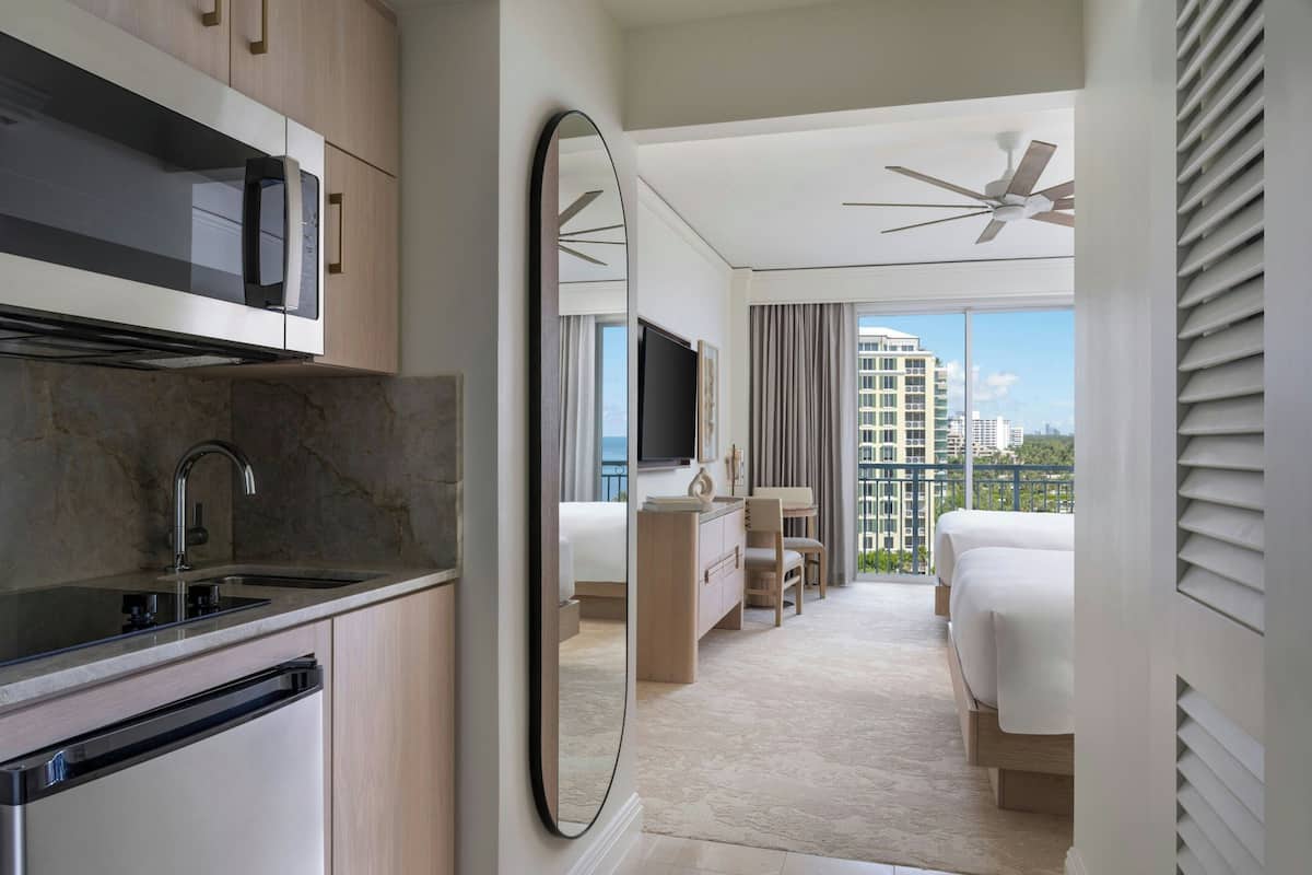 A modern hotel room with a kitchen area, a large mirror, a bed, a desk, and a view of the city.
