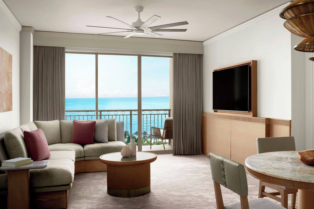 A modern hotel room with a curved sofa, a round coffee table, a dining table with chairs, a flat-screen TV mounted on the wall, and a balcony with a view of the ocean.