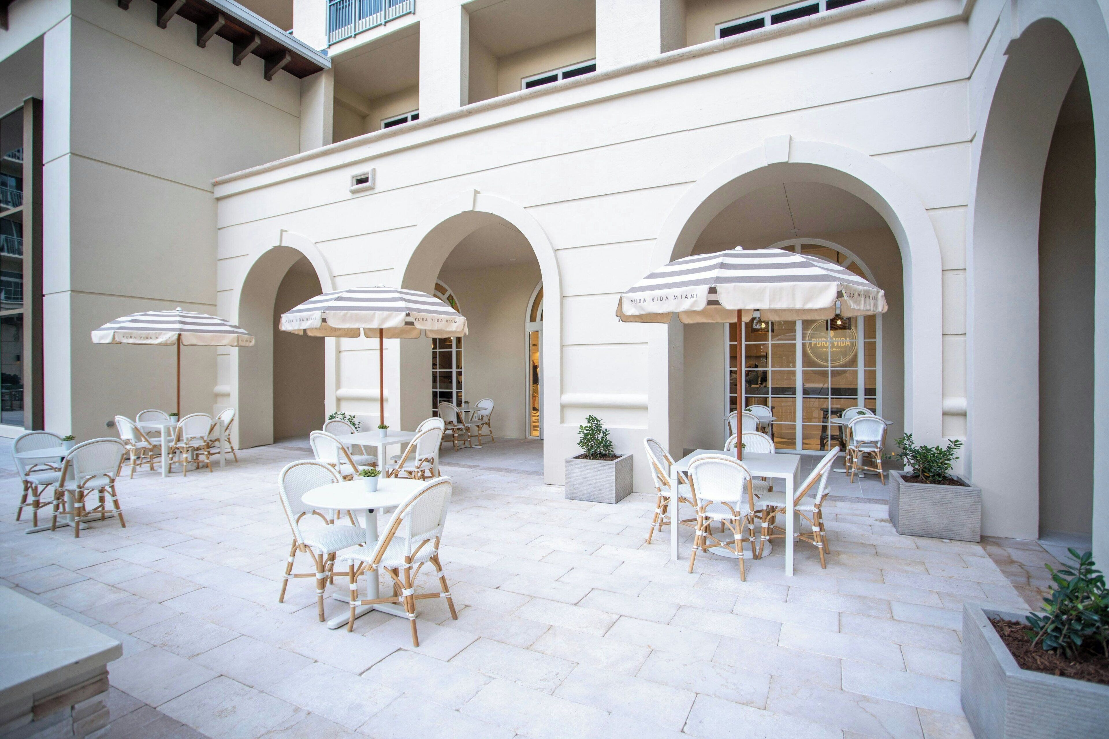 A courtyard with outdoor seating, white tables, and chairs, surrounded by arches and potted plants.