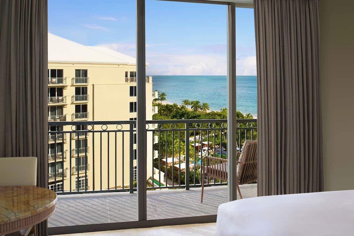 A hotel room with a large balcony, a view of the ocean, and a pool area with palm trees.