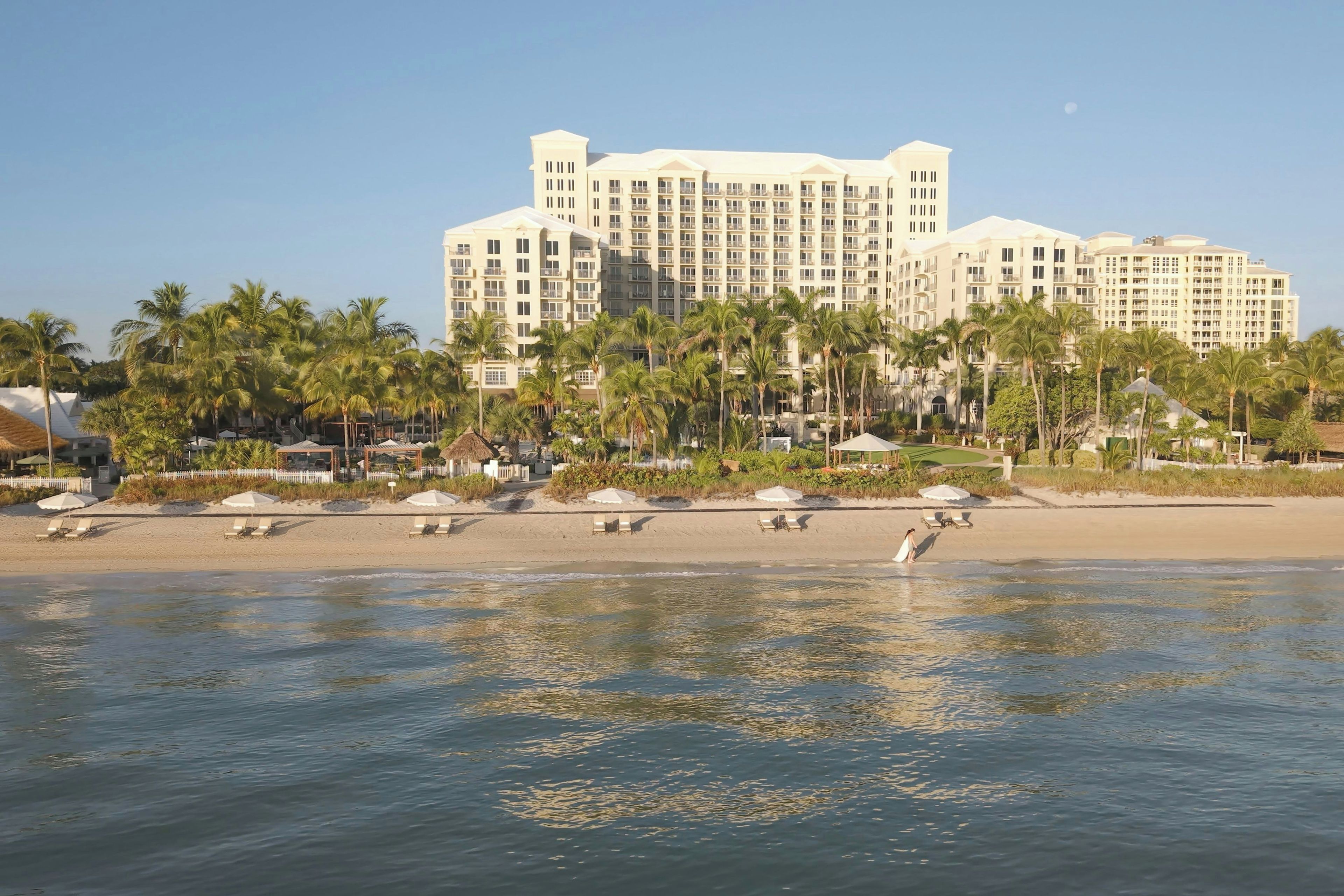 A beachfront hotel with multiple floors, palm trees, and a sandy beach.