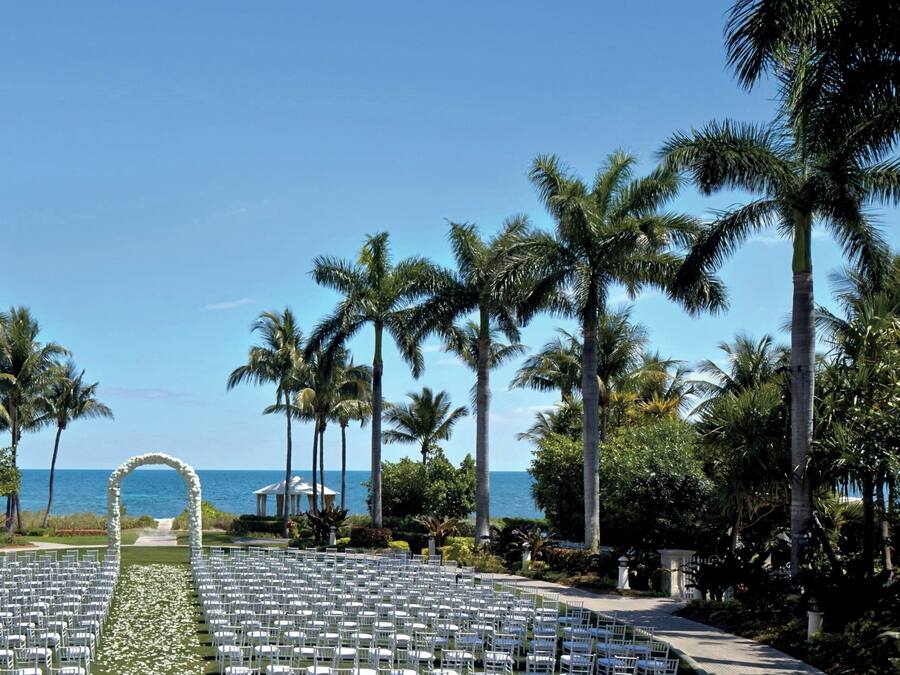 Eine Hochzeitseinrichtung am Strand mit Reihen weißer Stühle, einem Blumengang und Palmen.