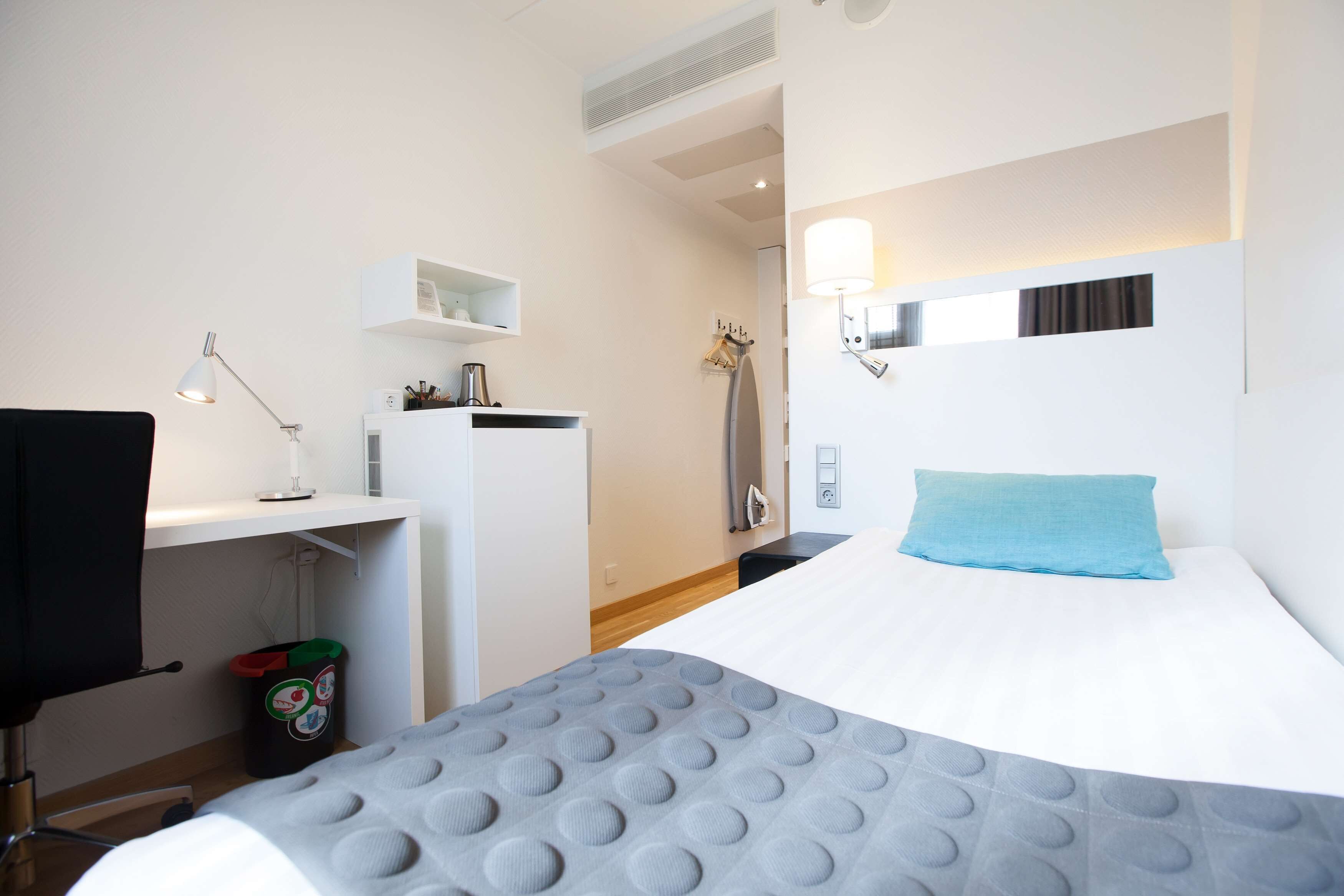 A hotel room with a bed, a desk, a chair, a trash bin, and a wall-mounted lamp.