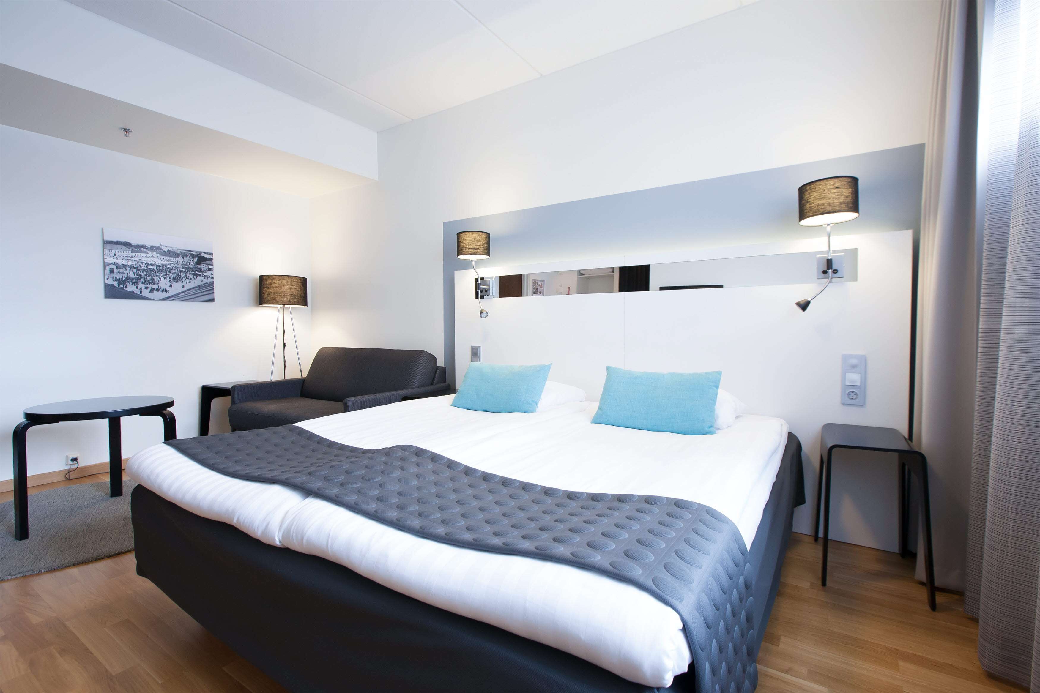 A modern hotel room with a large bed, two bedside lamps, a small table, and a chair.