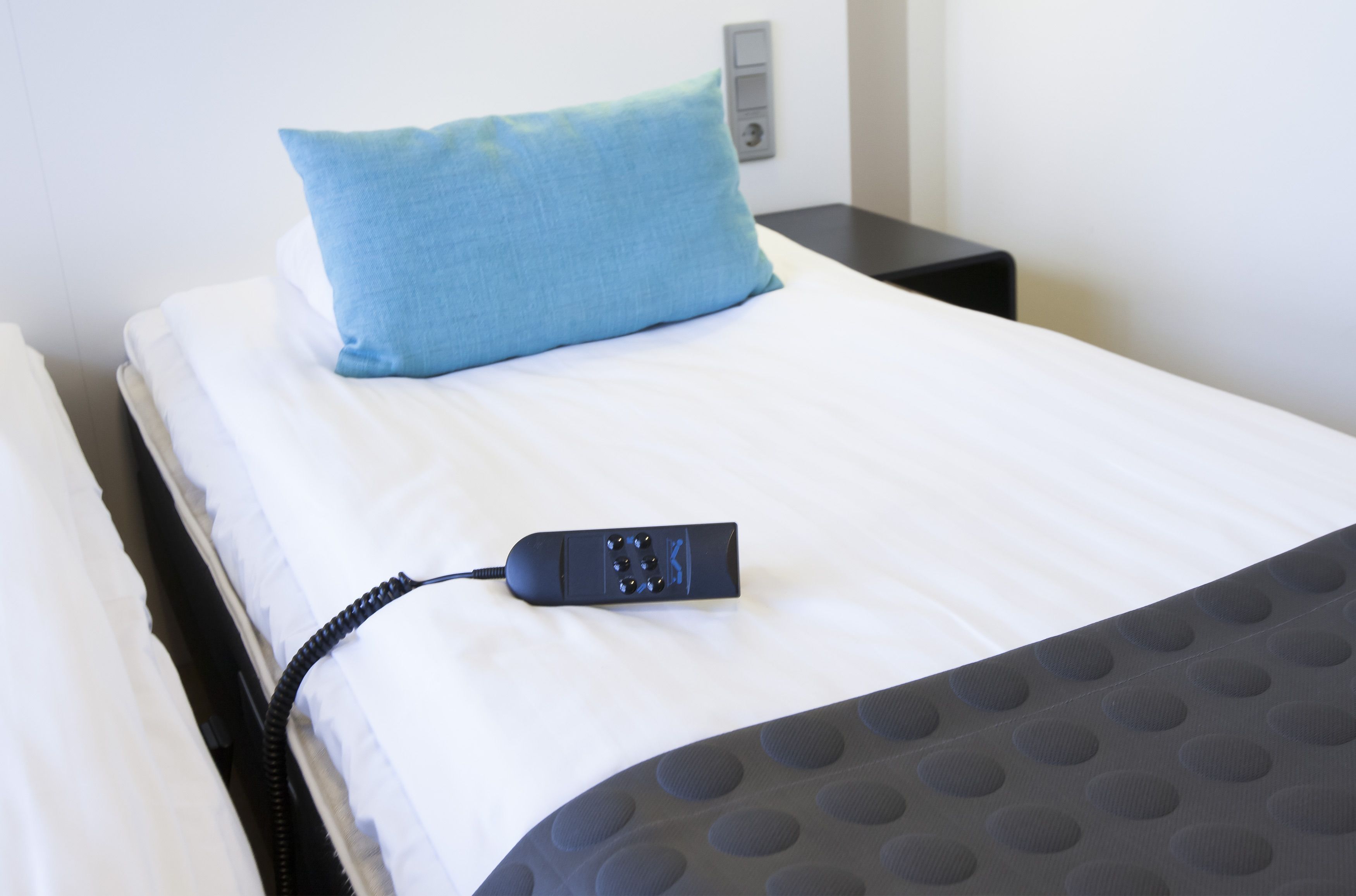 A bed with a blue pillow, a black remote control, and a gray patterned blanket.