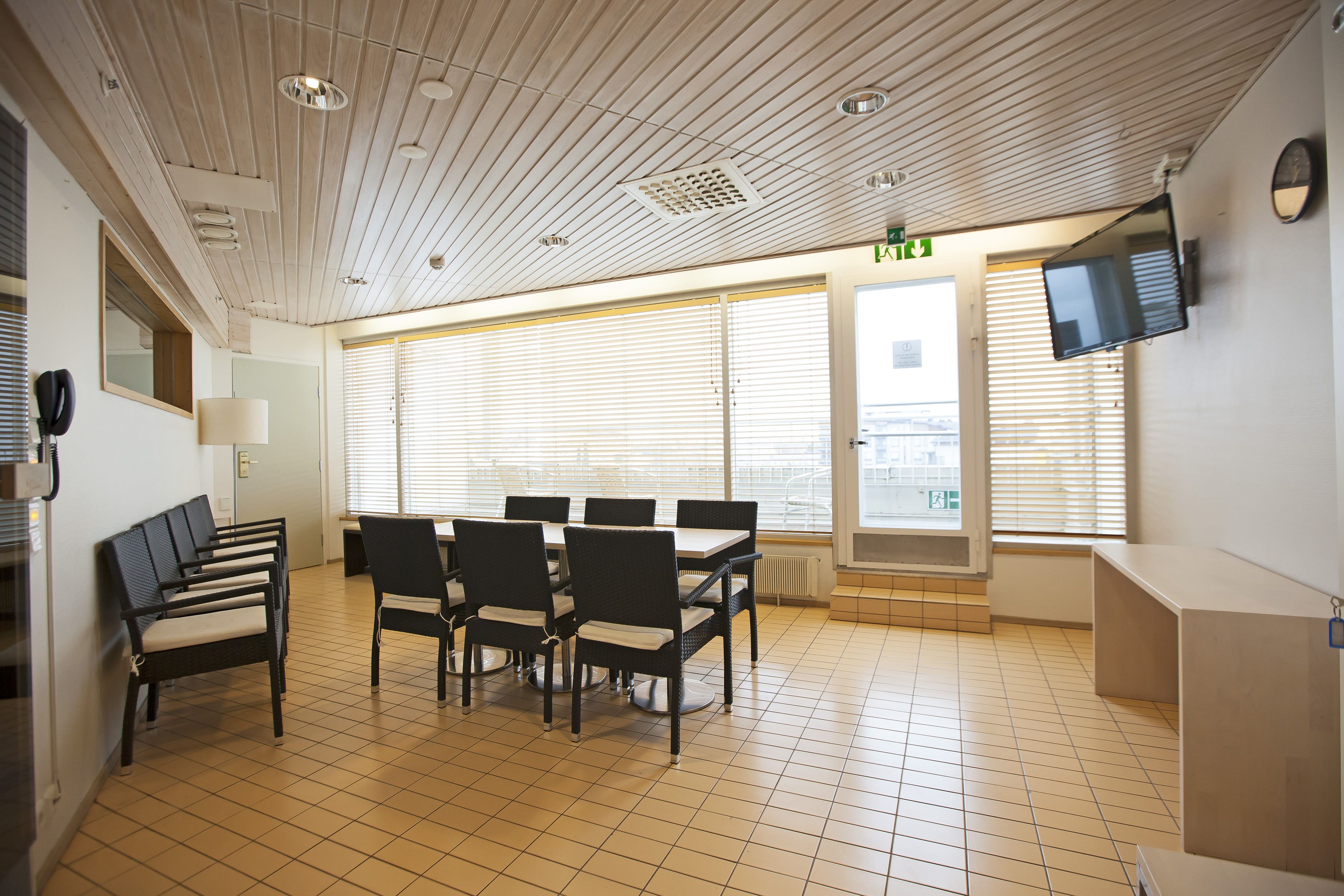 A conference room with a high ceiling, large windows with blinds, a flat-screen TV mounted on the wall, and a tiled floor.