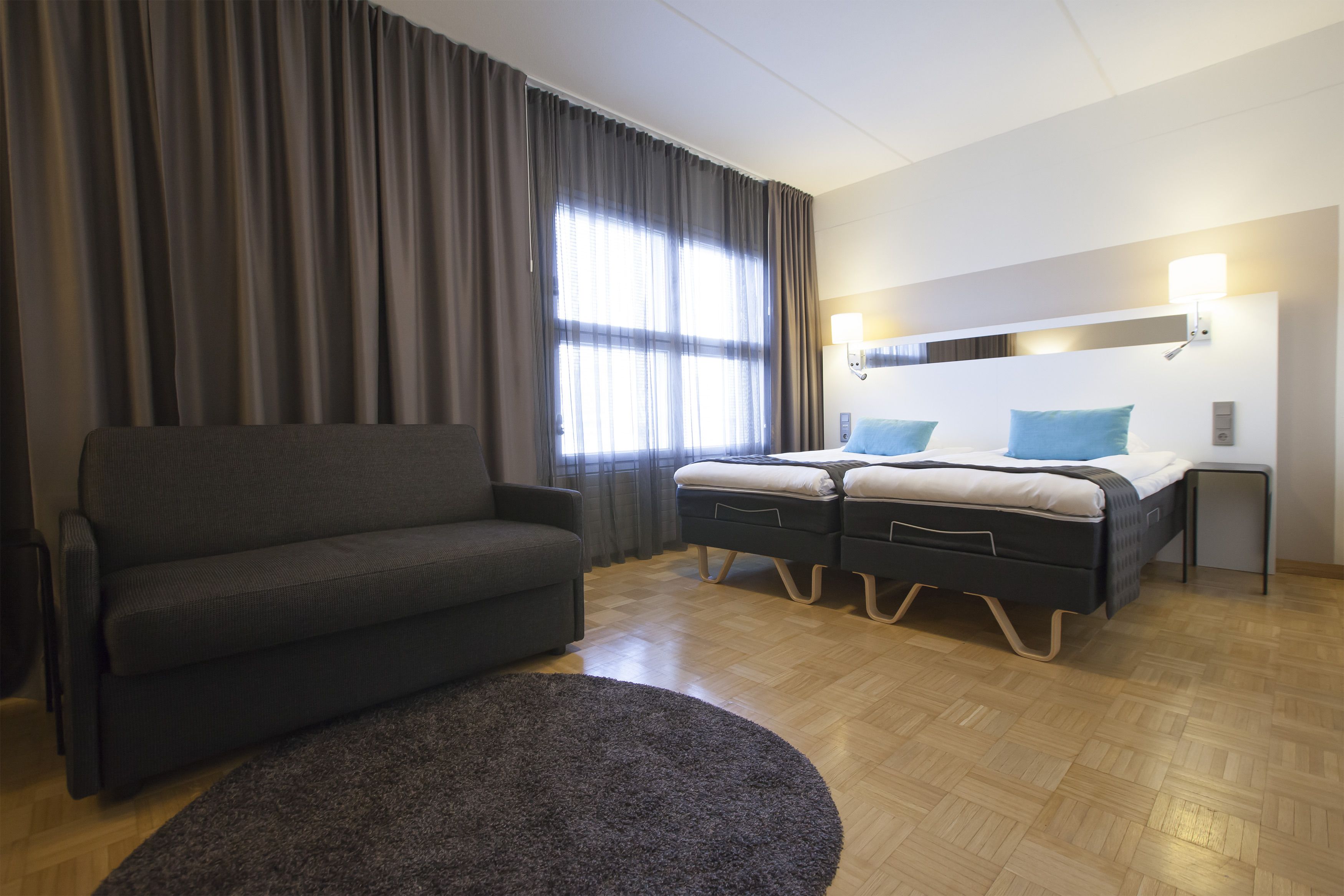 A modern hotel room with a sofa, a bed with two pillows, a bedside table, and a large window with curtains.