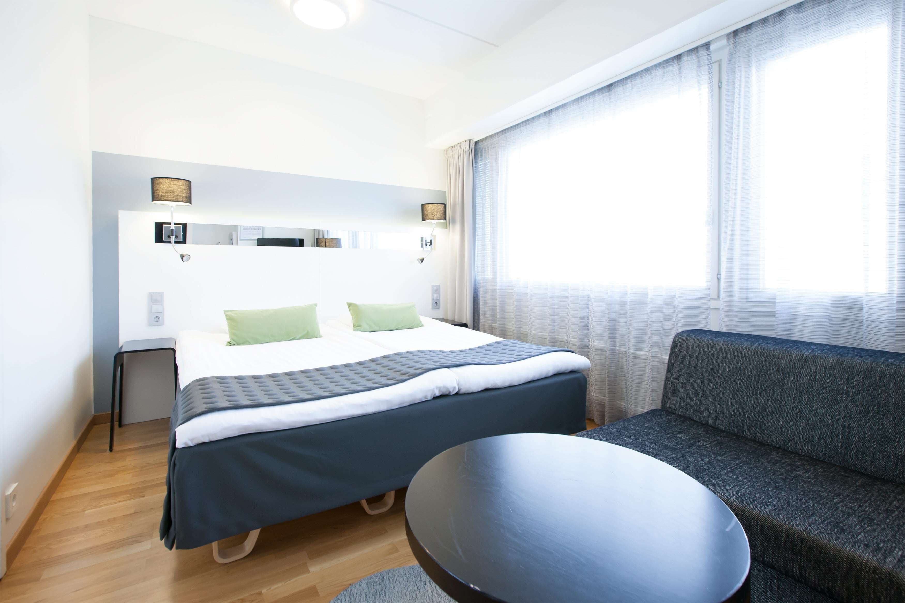 A modern hotel room with a bed, a sofa, a small table, and a window with curtains.