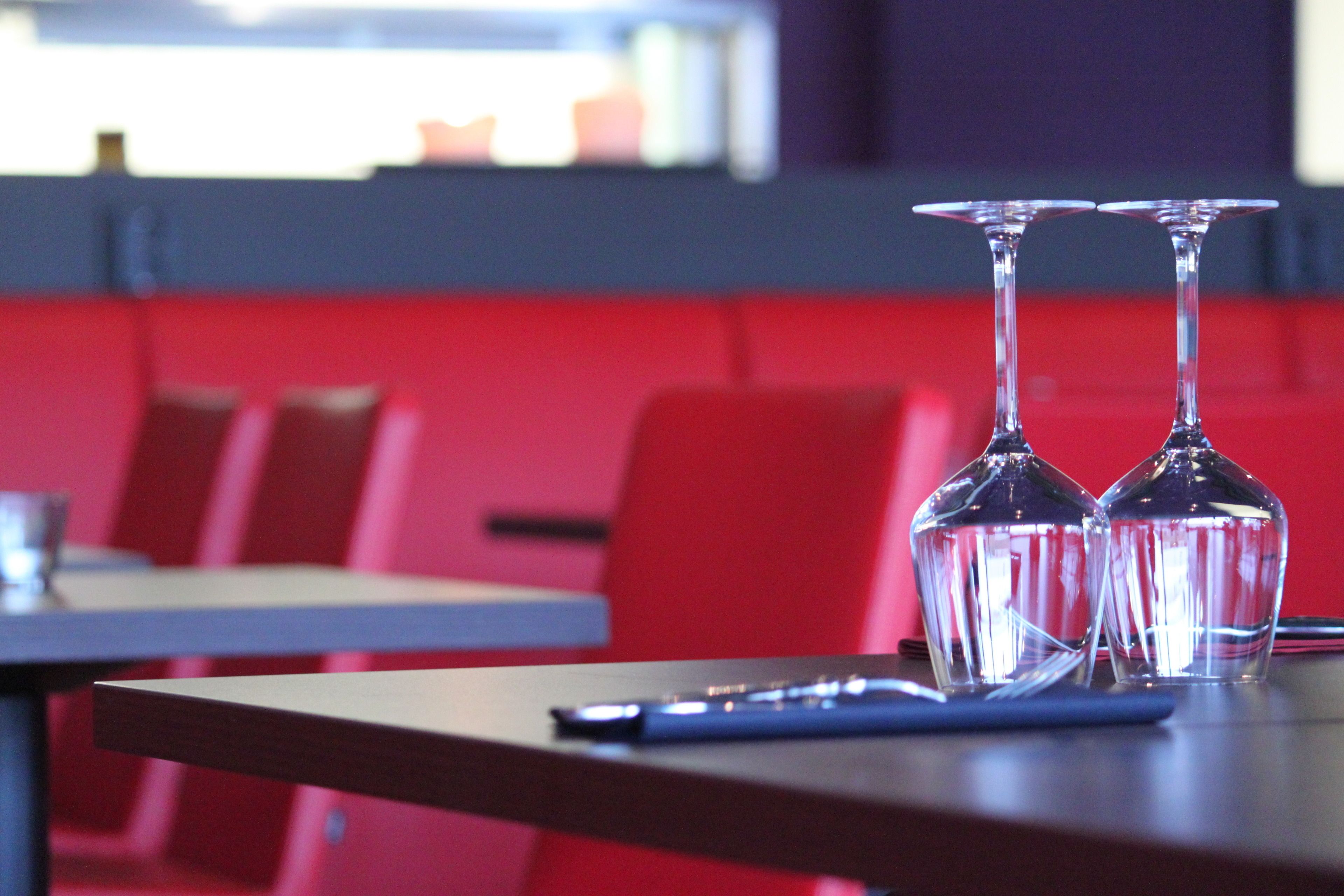 Une salle de restaurant avec deux verres à vin vides sur une table, des sièges rouges et une fenêtre en arrière-plan.