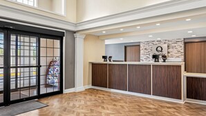 A hotel reception area with a herringbone wood floor, a reception desk with a 'Quality' sign, and a large glass door leading outside.