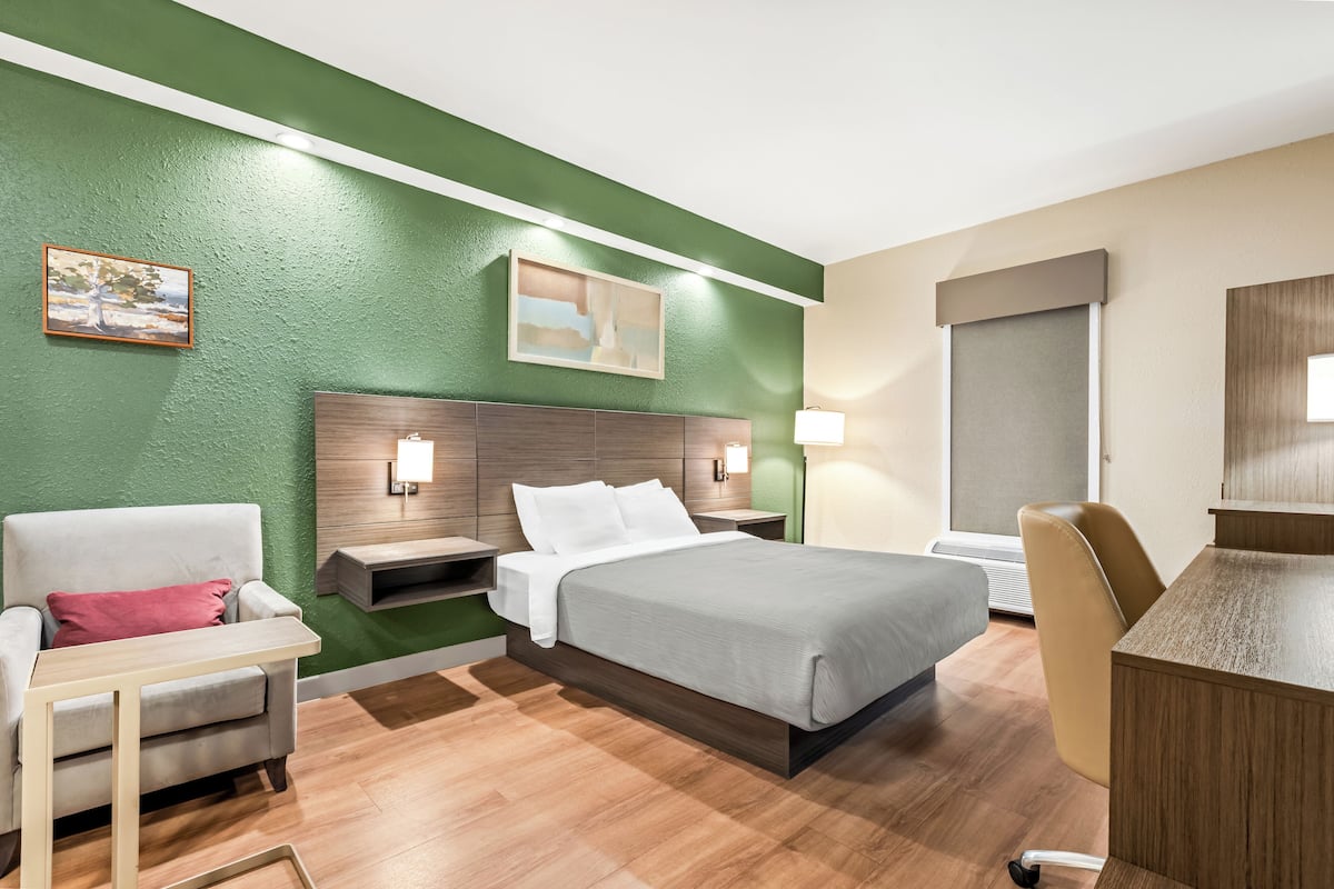 A hotel room with a bed, a desk, a chair, a nightstand, and a lamp.
