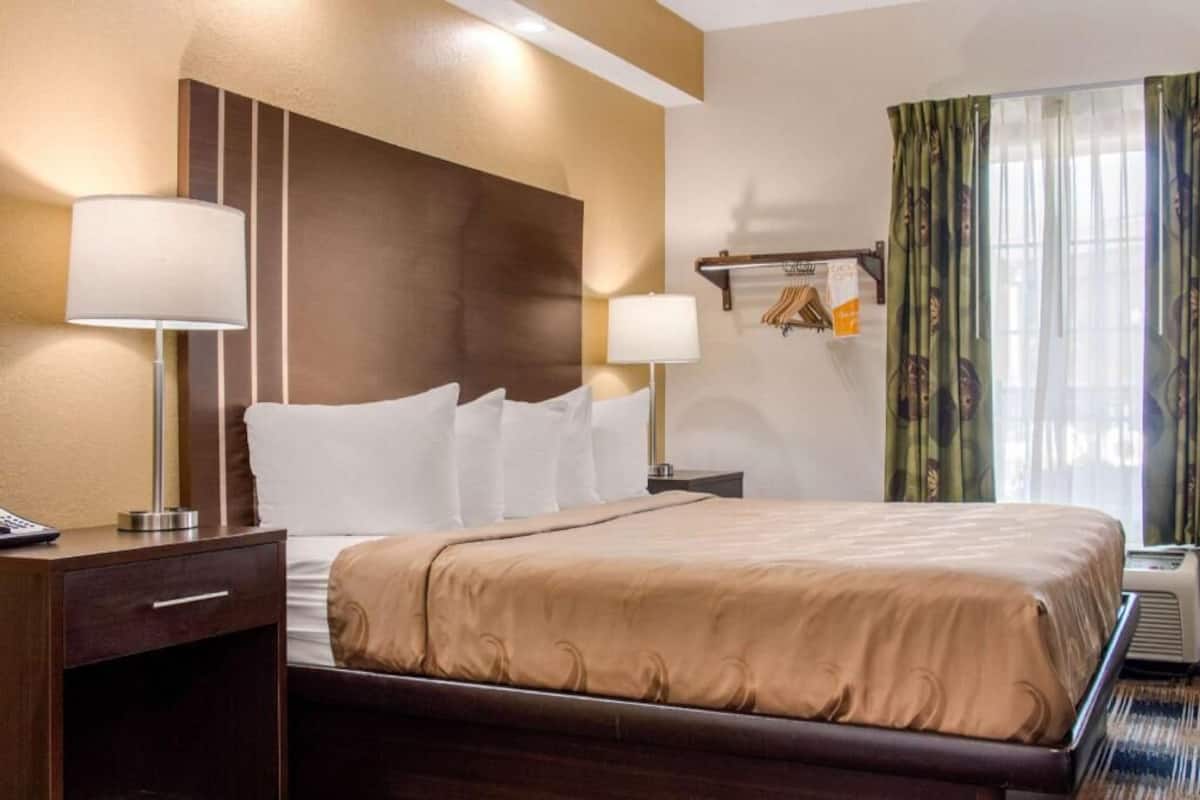 A hotel room with a large bed, two bedside lamps, a nightstand, a window with curtains, and a wall-mounted coat rack.