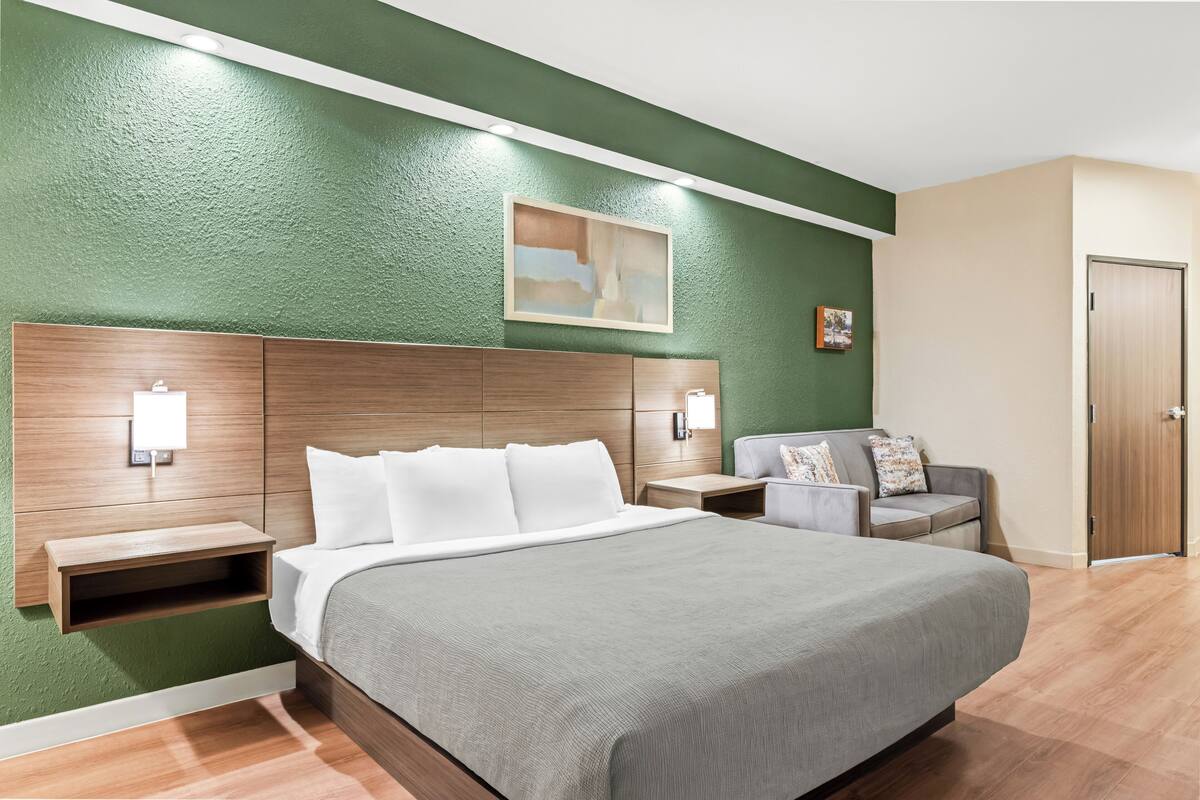 A hotel room with a large bed, a wooden headboard, a nightstand, a seating area with a sofa, and a door.