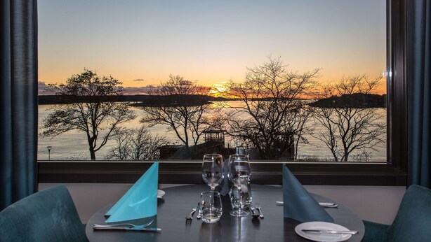 A dining table with a view of a sunset over a body of water, with trees and a gazebo in the background.