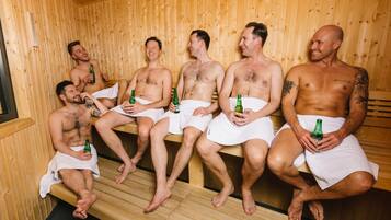 Una sauna con paredes y bancos de madera, donde cinco hombres están sentados, cada uno sosteniendo una botella.