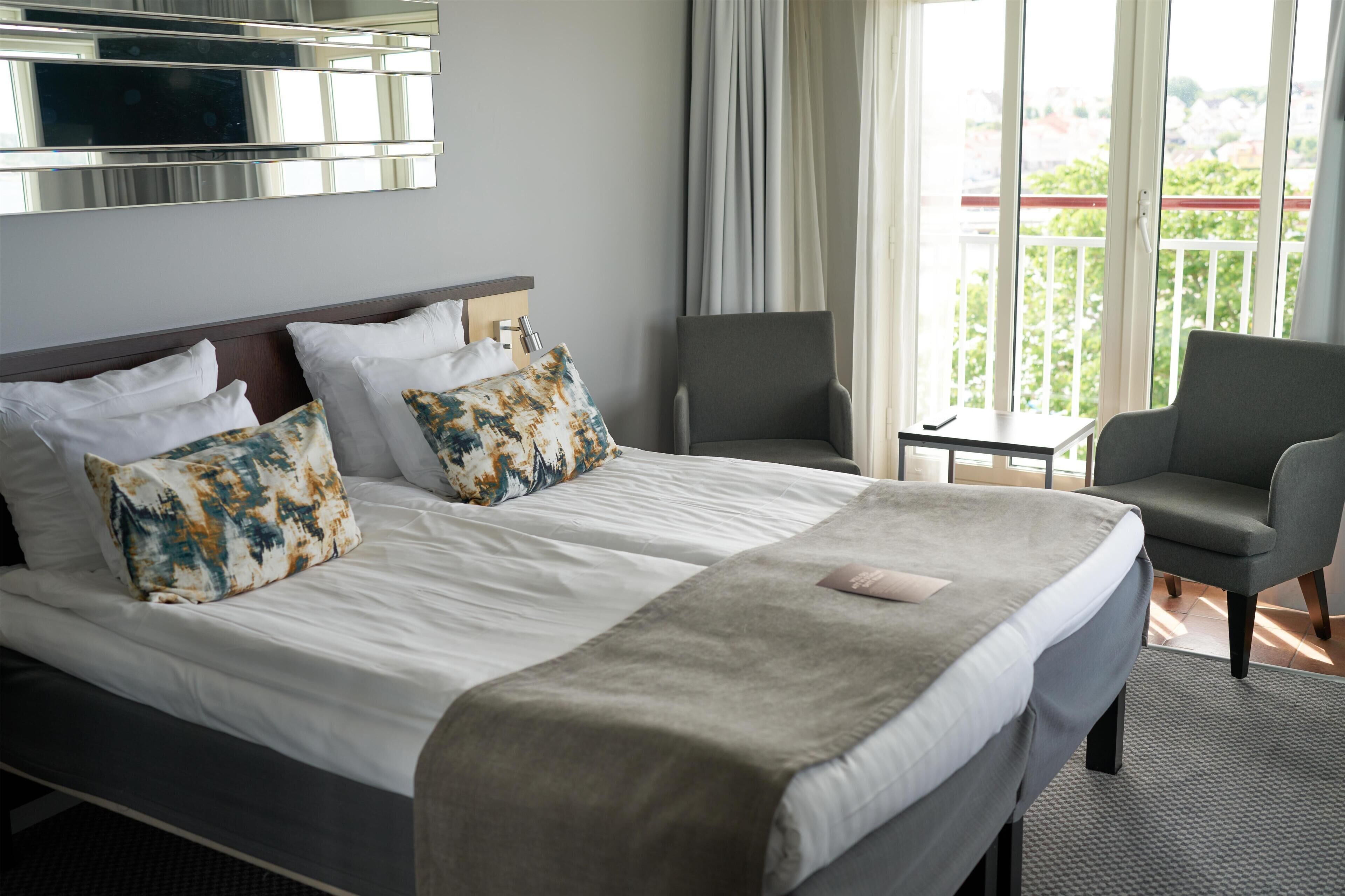 Habitación de hotel con cama, dos sillas, una mesita, televisor y balcón con barandilla.