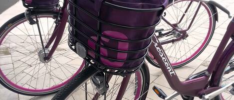 A bicycle with a purple basket and a pink frame parked on a tiled pavement.