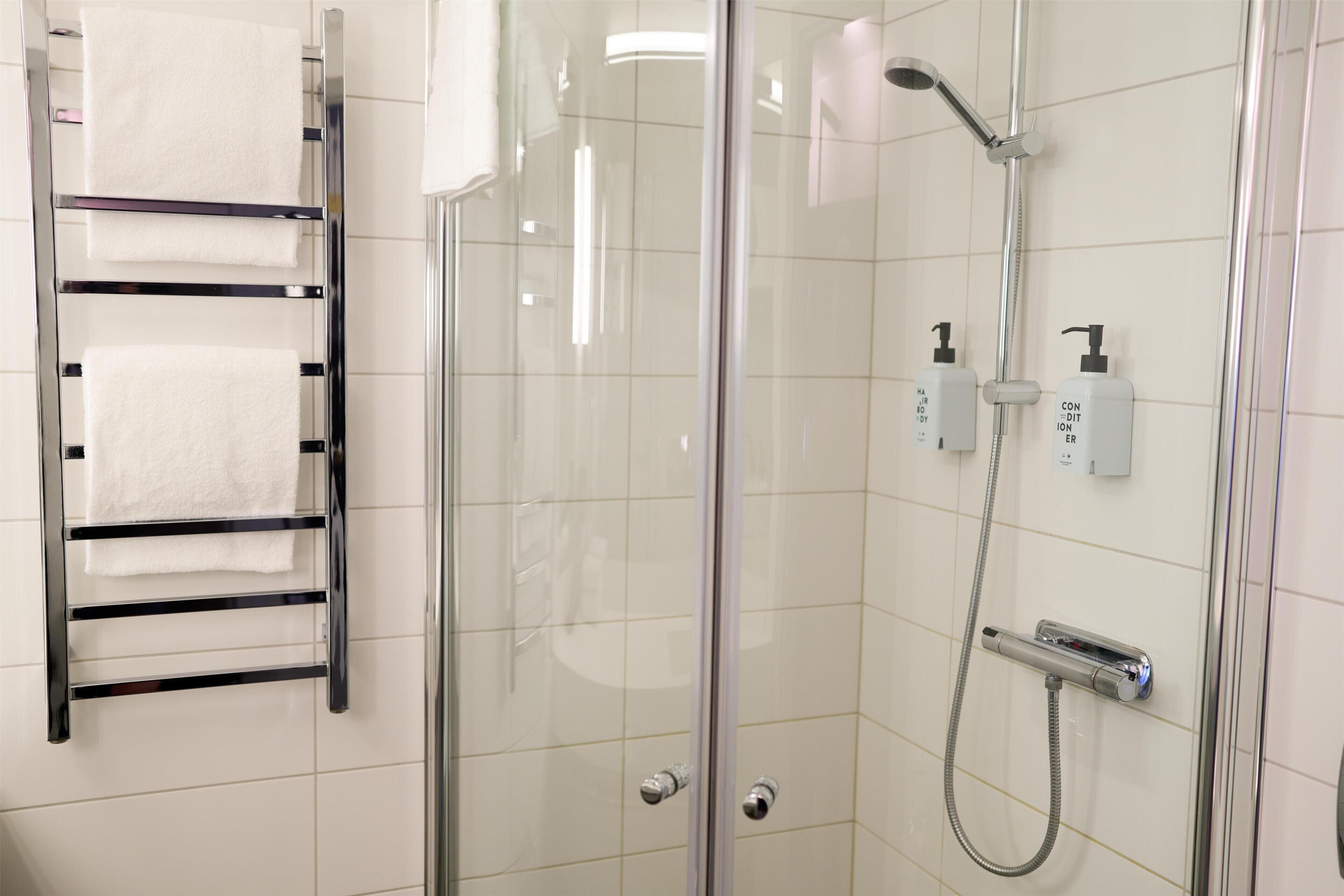 A glass-enclosed shower with a handheld showerhead, a towel rack with white towels, and two bottles of soap on the wall.
