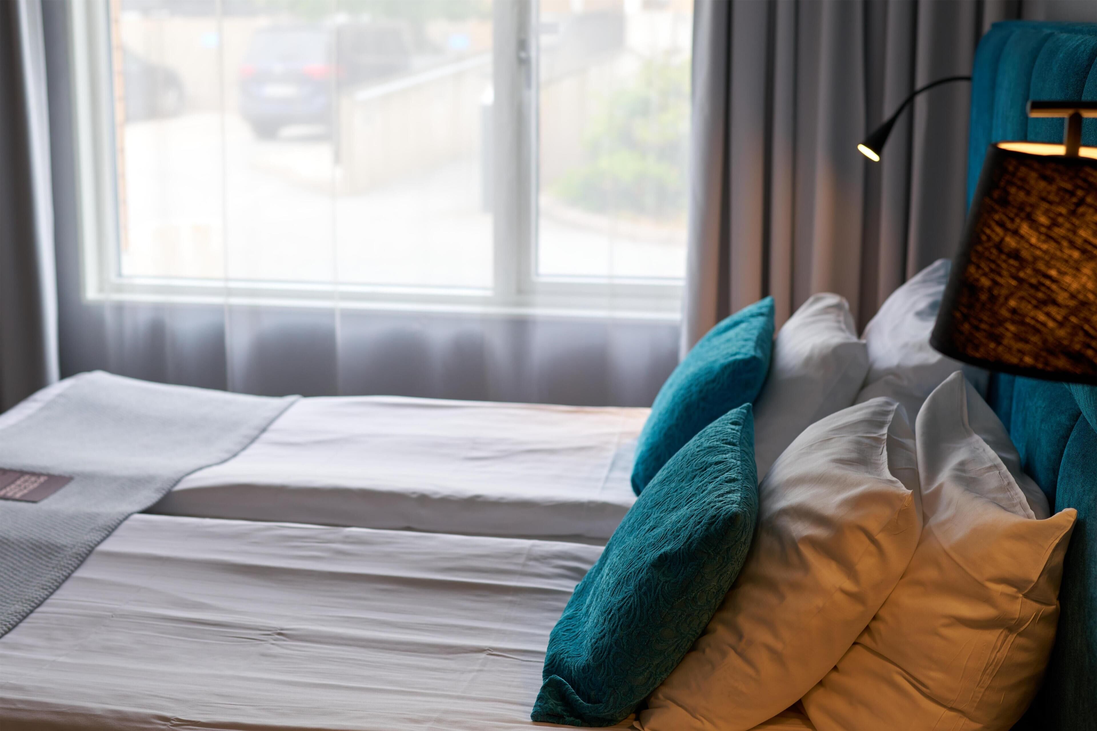 Una cama con sábanas blancas y almohadas verde azulado y beige, una lámpara con pantalla texturizada, y una ventana con vista a la calle y coches aparcados.