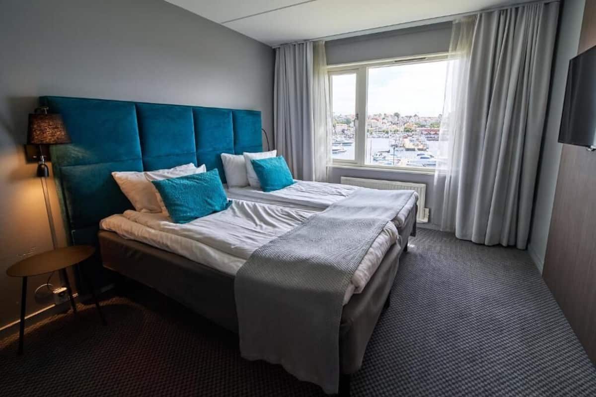 Habitación de hotel con una cama grande, una mesita de noche, una lámpara y vistas a la ciudad a través de la ventana.