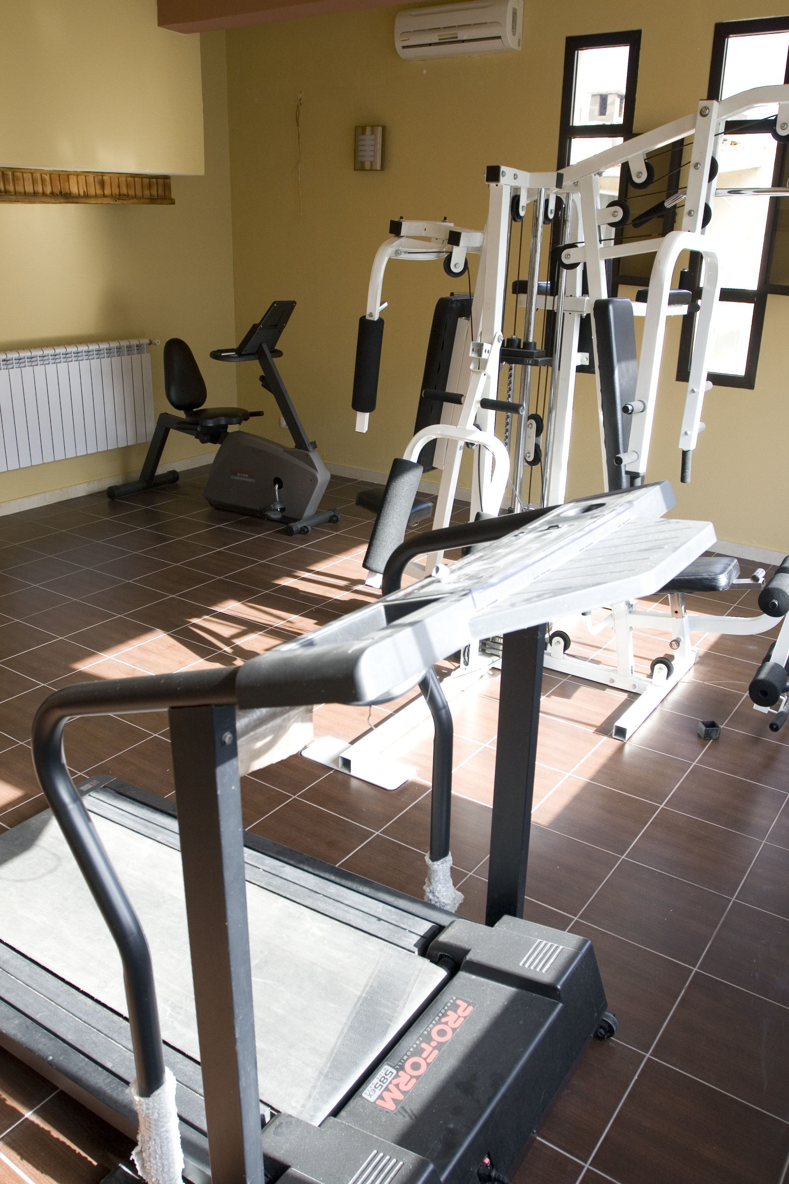 A gym with various exercise equipment, including treadmills and weight machines, in a room with tiled flooring and large windows.