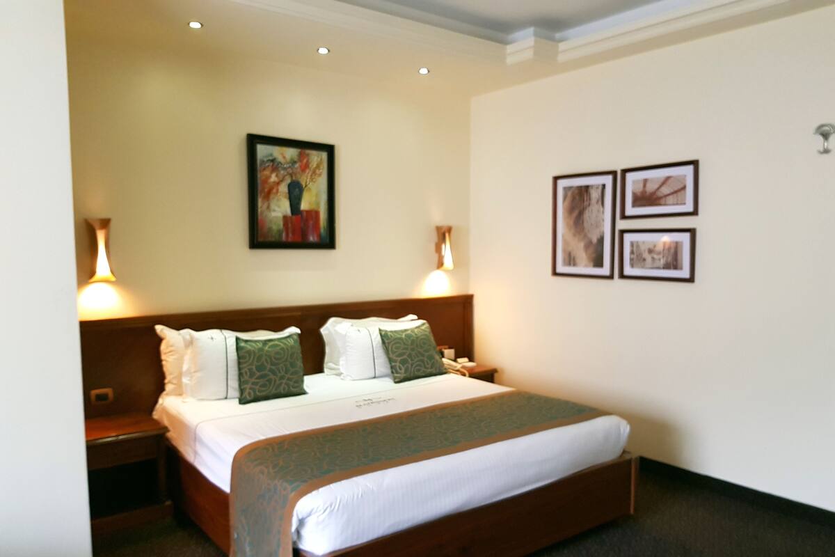 A hotel room with a large bed, two bedside tables, a nightstand, a painting on the wall, and two framed pictures.
