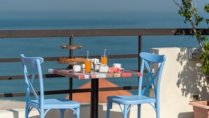 Une table avec un plateau de desserts, des tasses et un verre de jus d’orange, le tout disposé devant une vue sur la mer.
