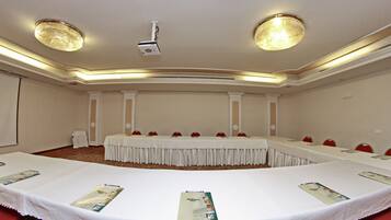A conference room with a long table, red chairs, white tablecloths, and a projector on the ceiling.