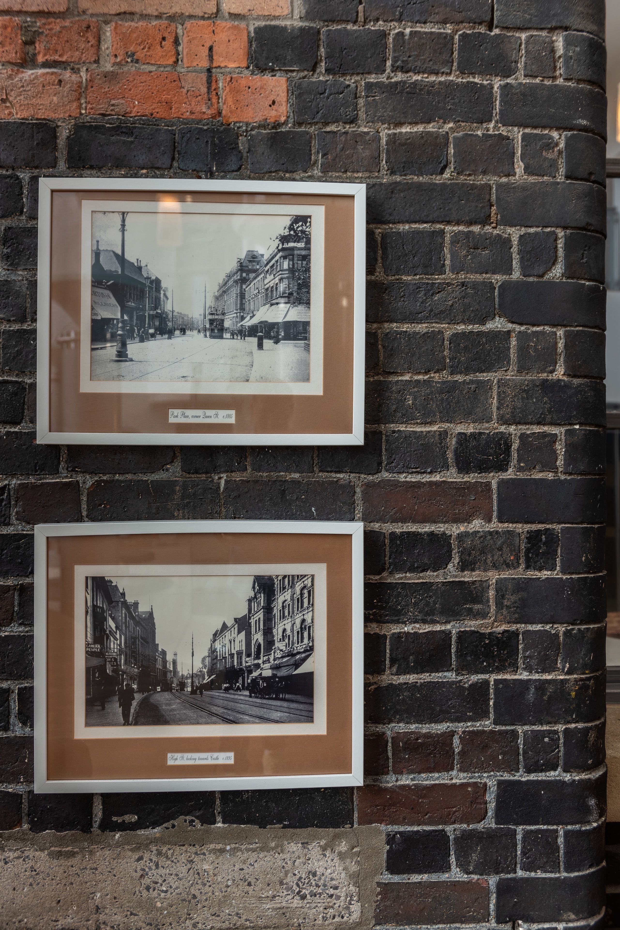 Två inramade svartvita fotografier som föreställer en gatuscen mot en mur av tegelstenar.