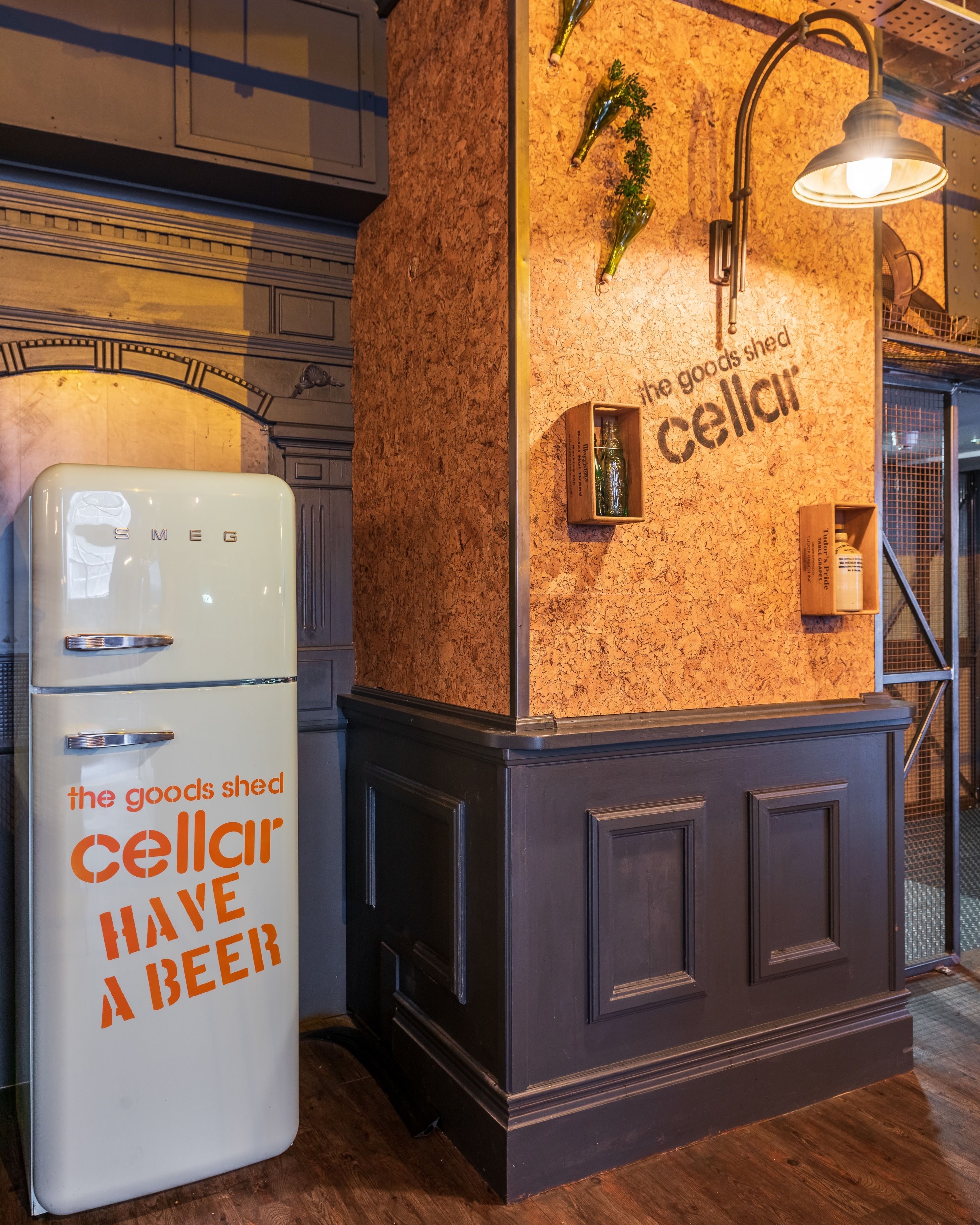 Una cocina con un refrigerador blanco, un mueble de madera y una pared con un tablón de corcho mostrando objetos y texto.