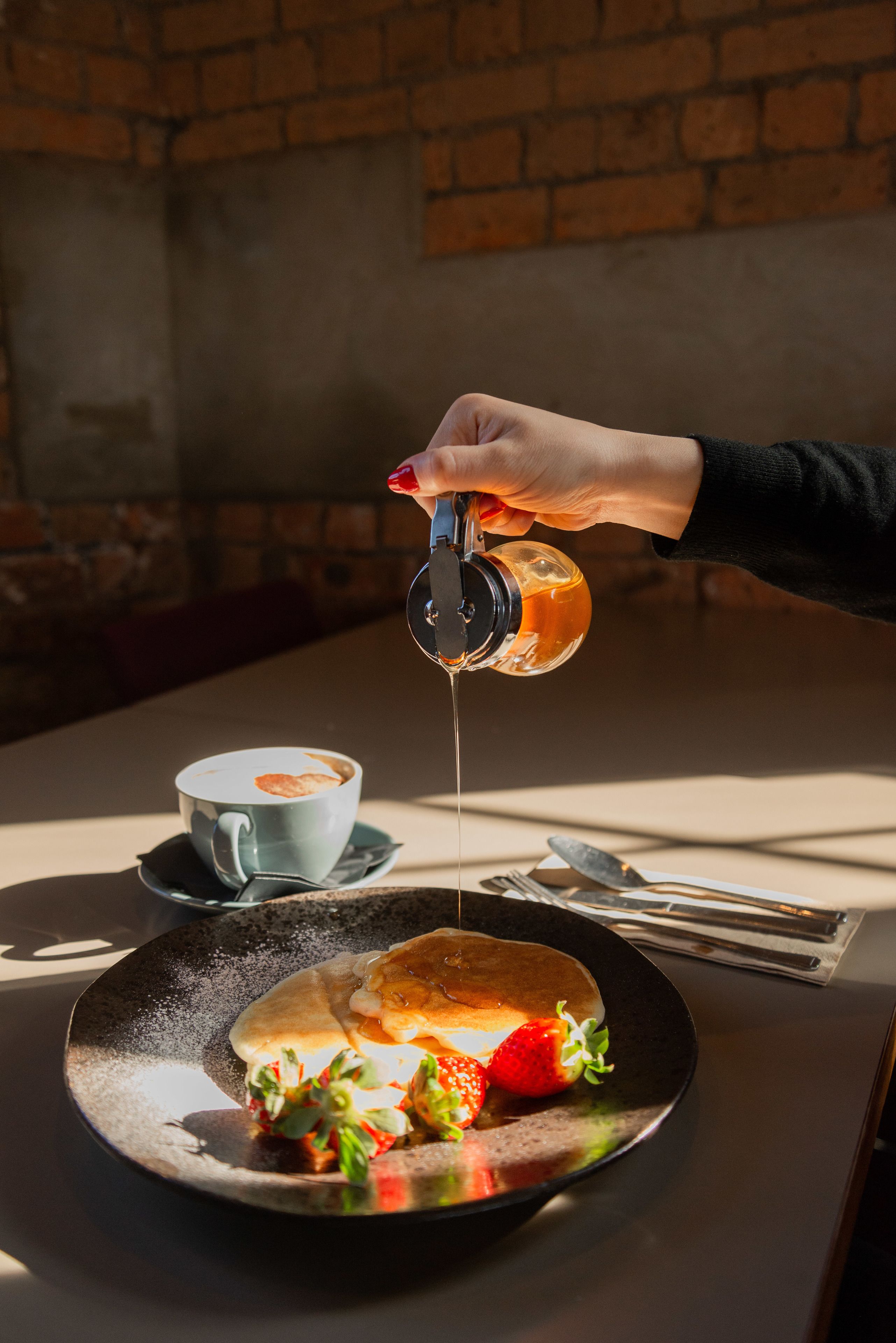 A hand pouring syrup onto a plate of food with a cup of coffee in the background.