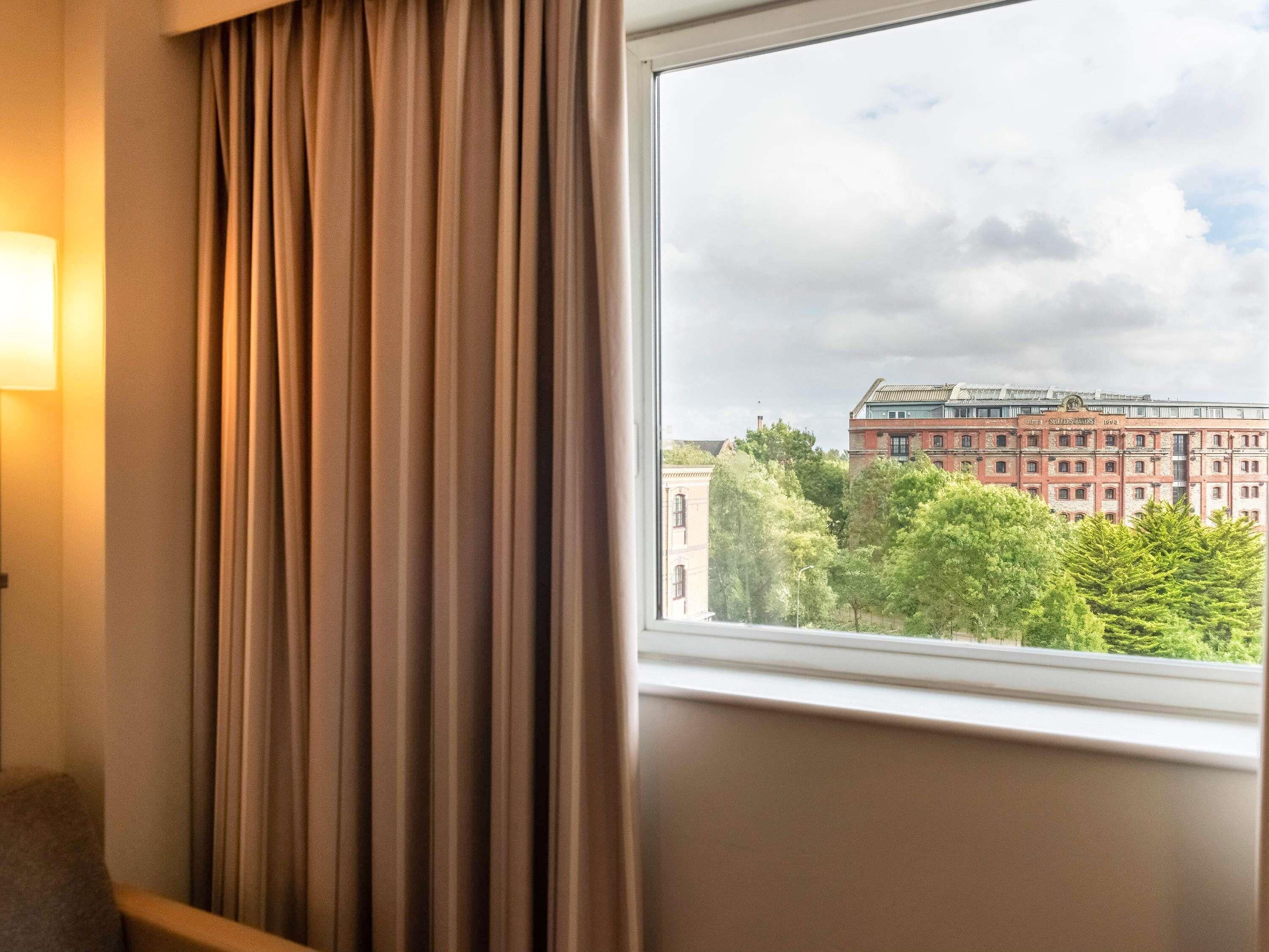 Hotelkamer met raam en uitzicht op een gebouw met rode gevel en bomen.