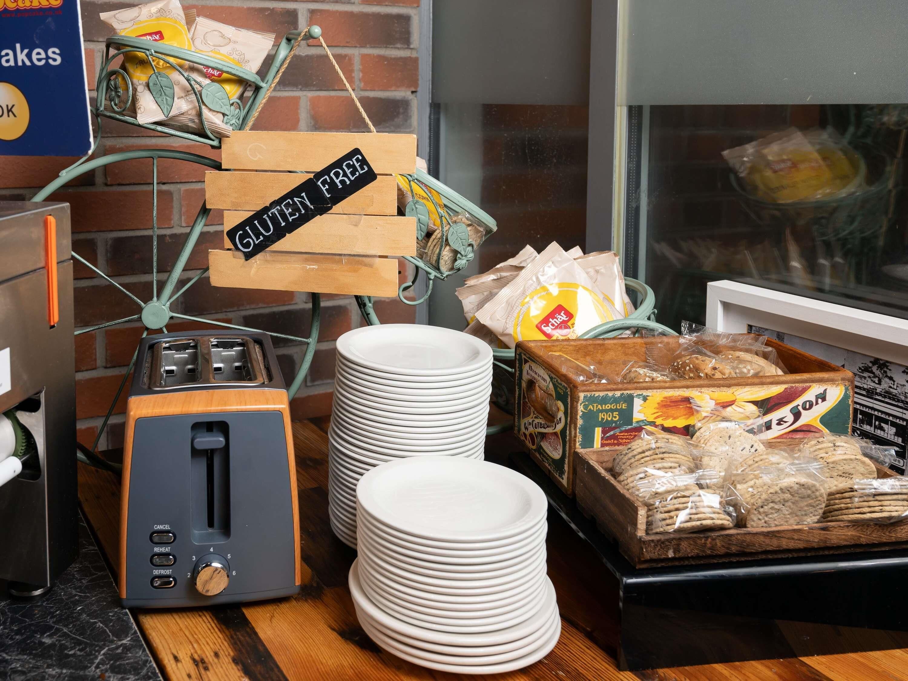 Una cocina con tostadora, pilas de platos blancos y productos etiquetados como sin gluten.