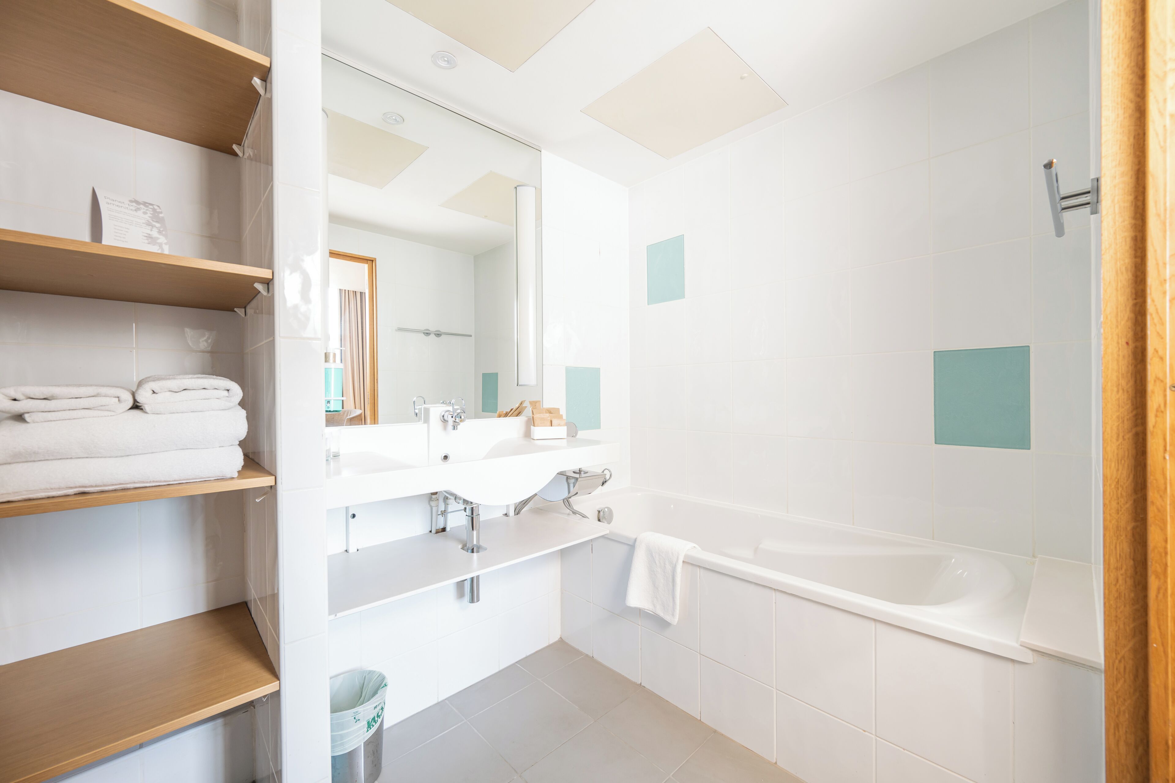 A modern bathroom with a bathtub, a sink, and a mirror. There are wooden shelves with towels and a trash bin.