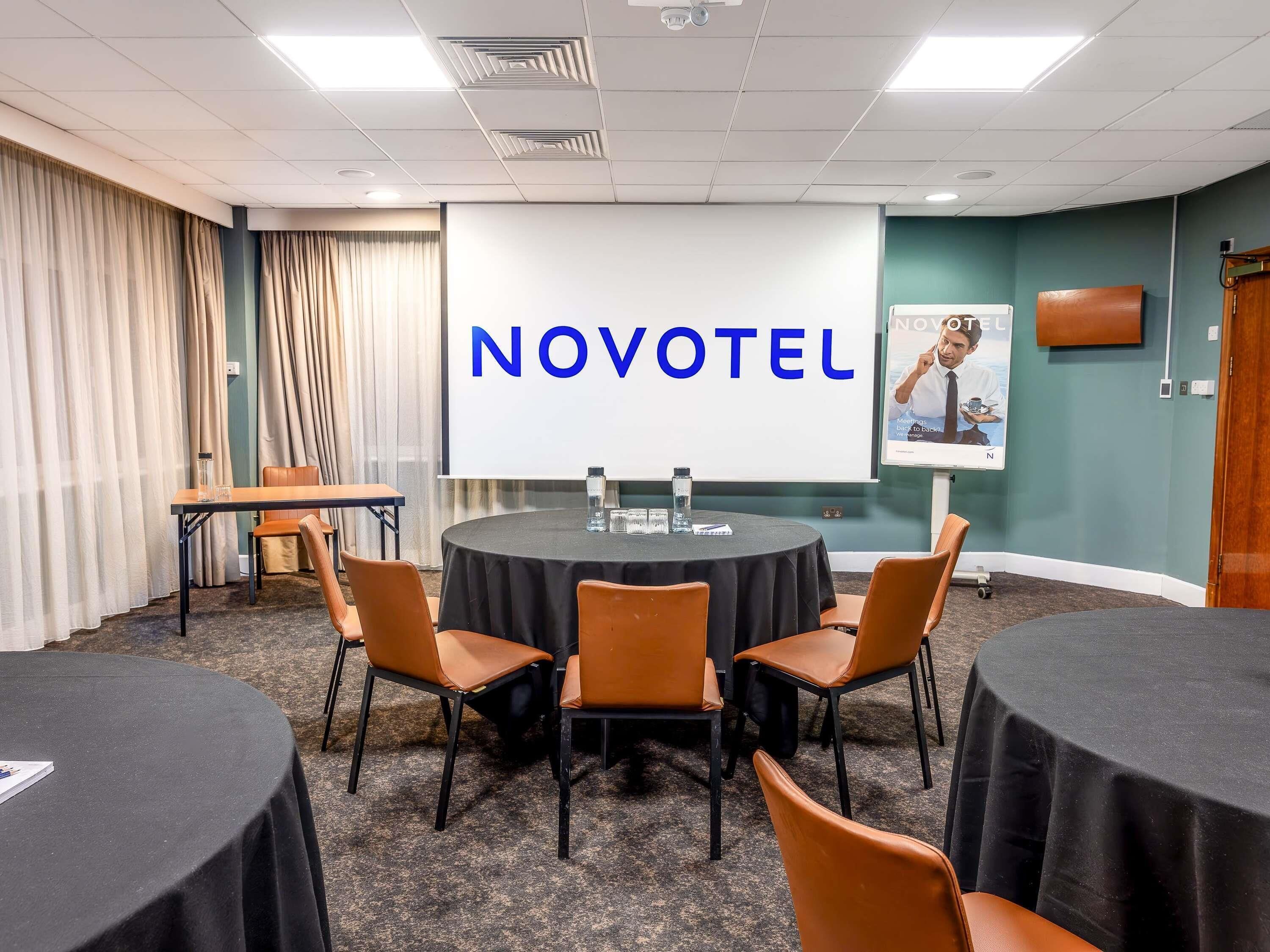 A conference room with a large screen displaying 'NOVOTEL,' a round table with chairs, and a smaller table with a laptop.