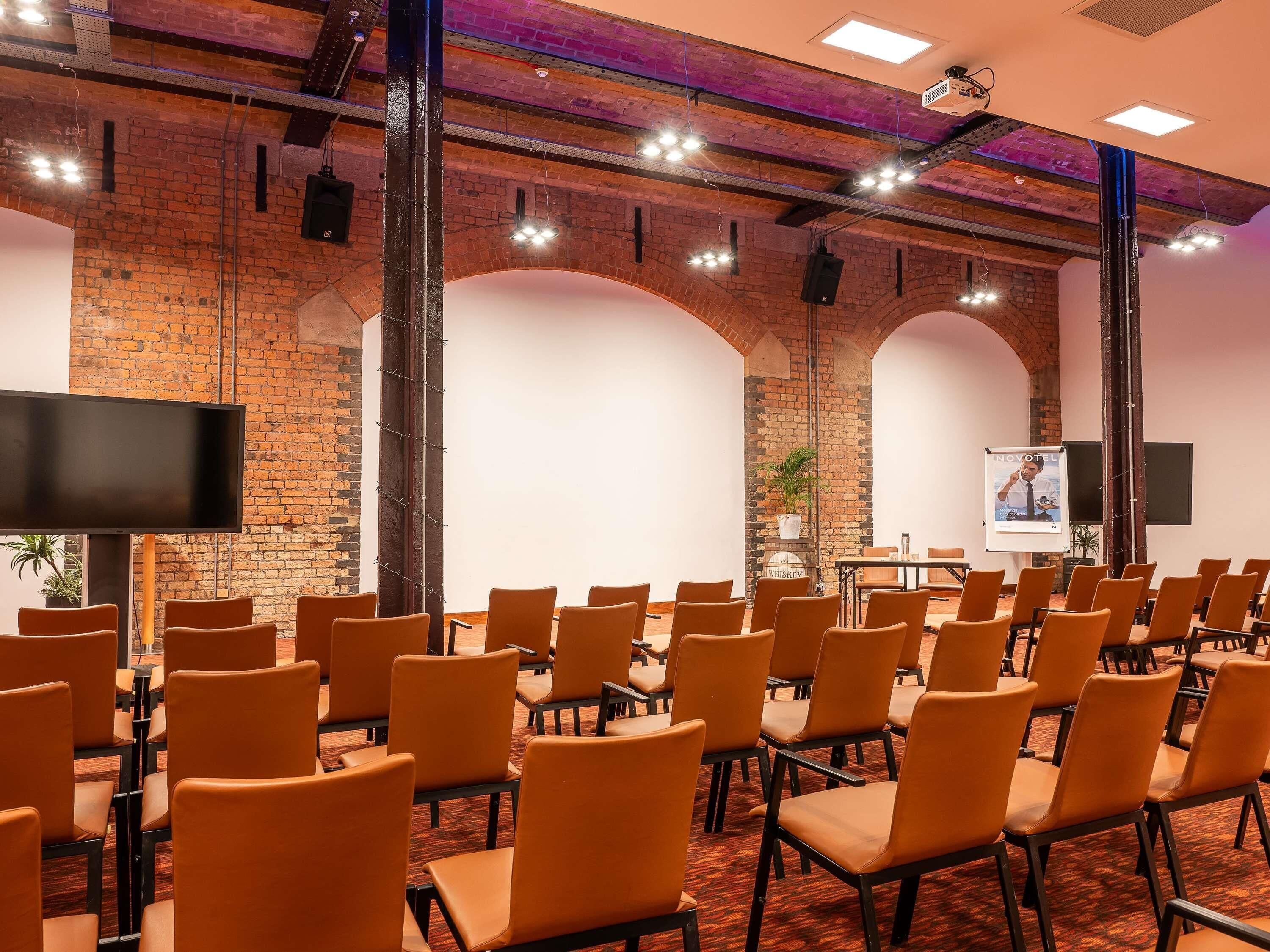 Une salle de conférence avec des rangées de chaises, un écran de projection et un mur en briques.