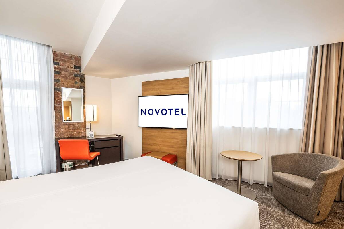 A hotel room with a large bed, a desk with a chair, a small round table, and a TV displaying "NOVOTEL".