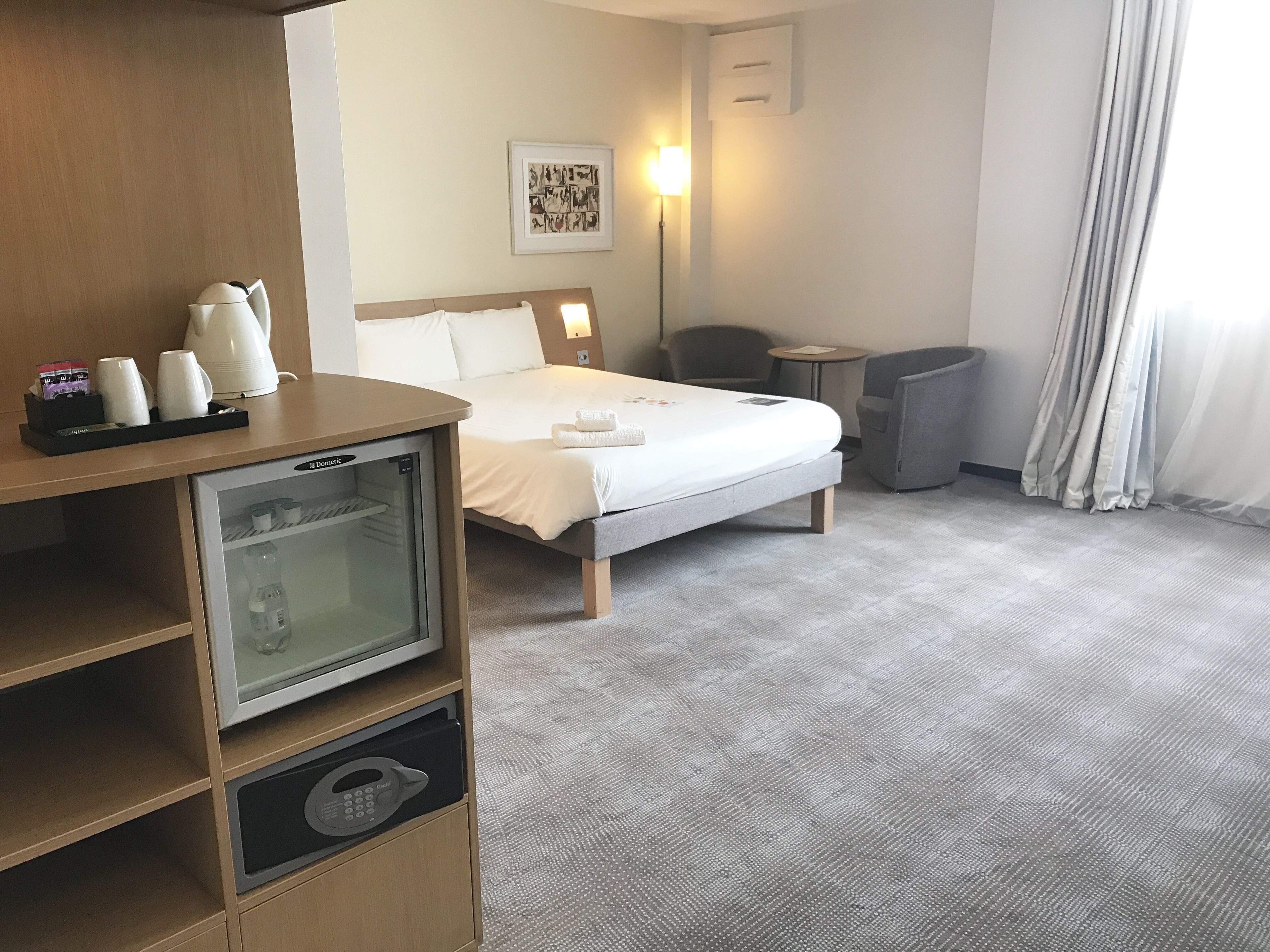 A hotel room with a bed, a minibar, a seating area, and a wall-mounted picture.