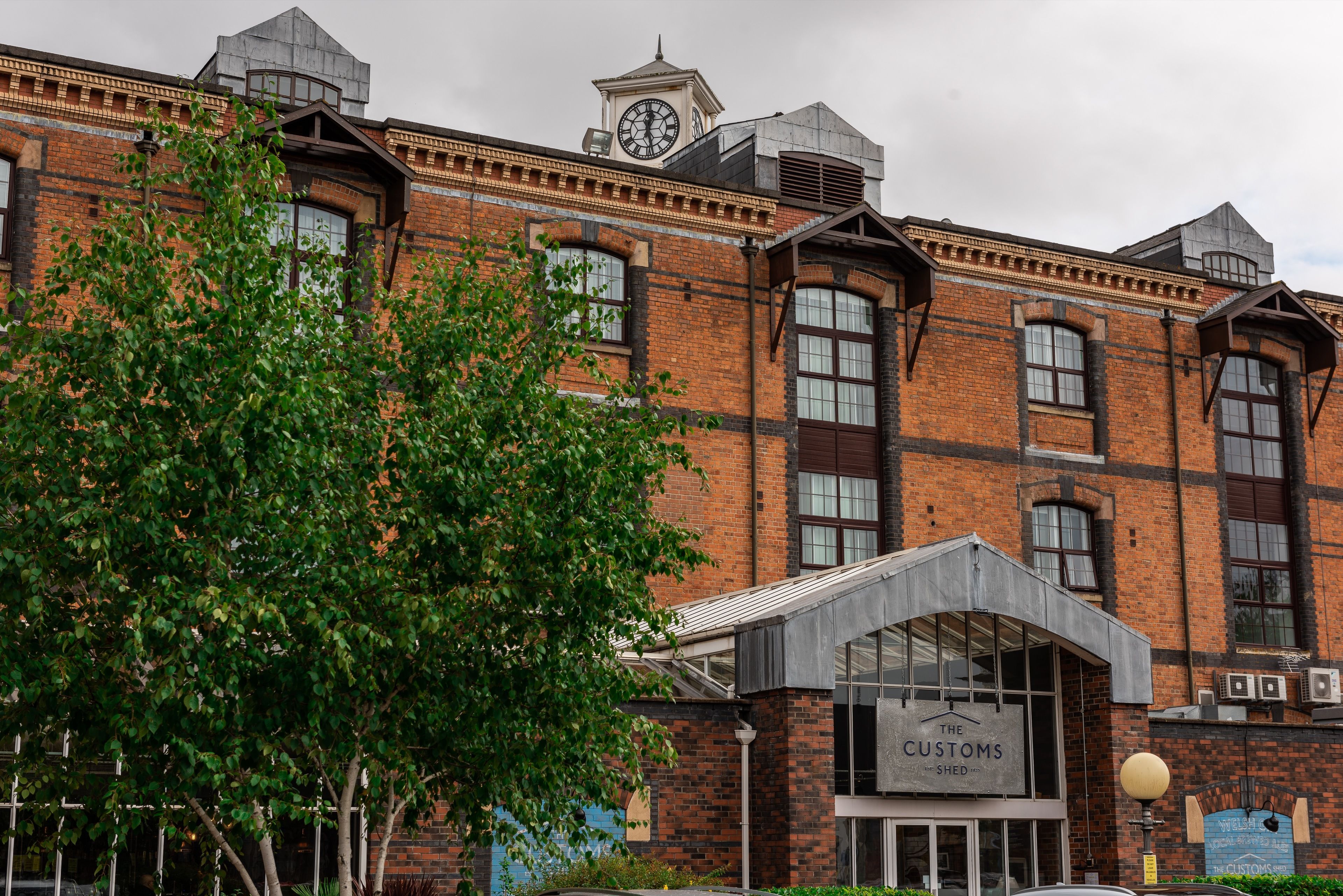 En historisk bygning med et tårn og et ur, store vinduer samt et skilt, der siger "The Customs Shed".