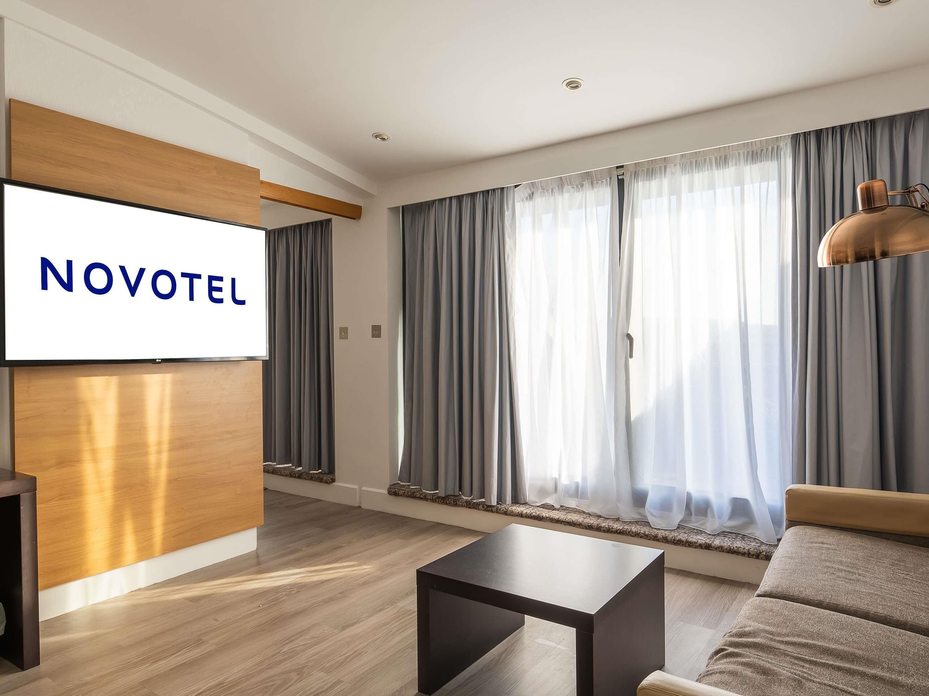 Ein modernes Hotelzimmer mit einem Flachbildfernseher, der 'NOVOTEL' zeigt, einem hölzernen Empfangsschreibtisch, einer Couch, einem Couchtisch und großen Fenstern mit Vorhängen.