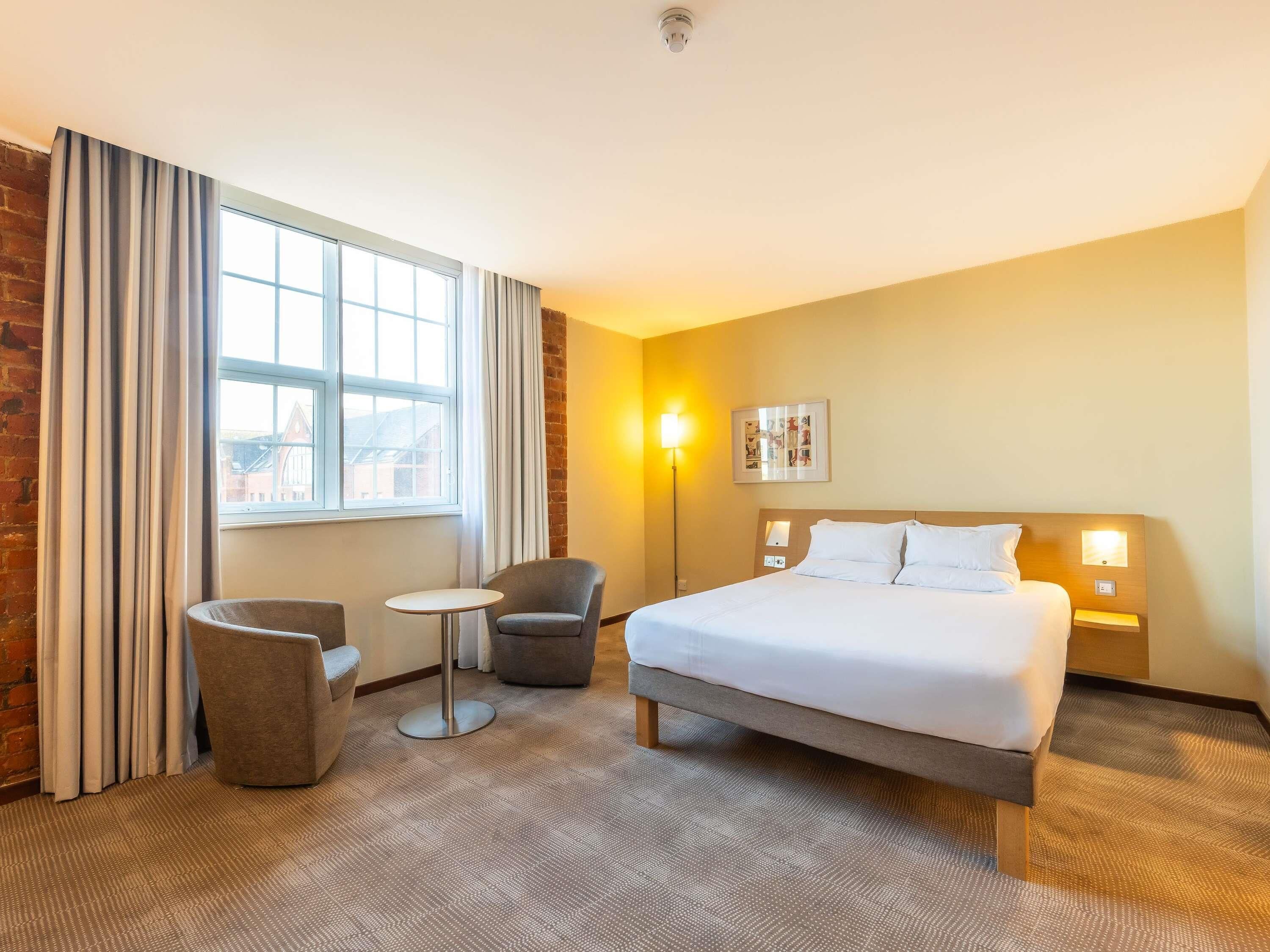 A hotel room with a large bed, a small round table, two armchairs, a window with curtains, and a wall-mounted light fixture.