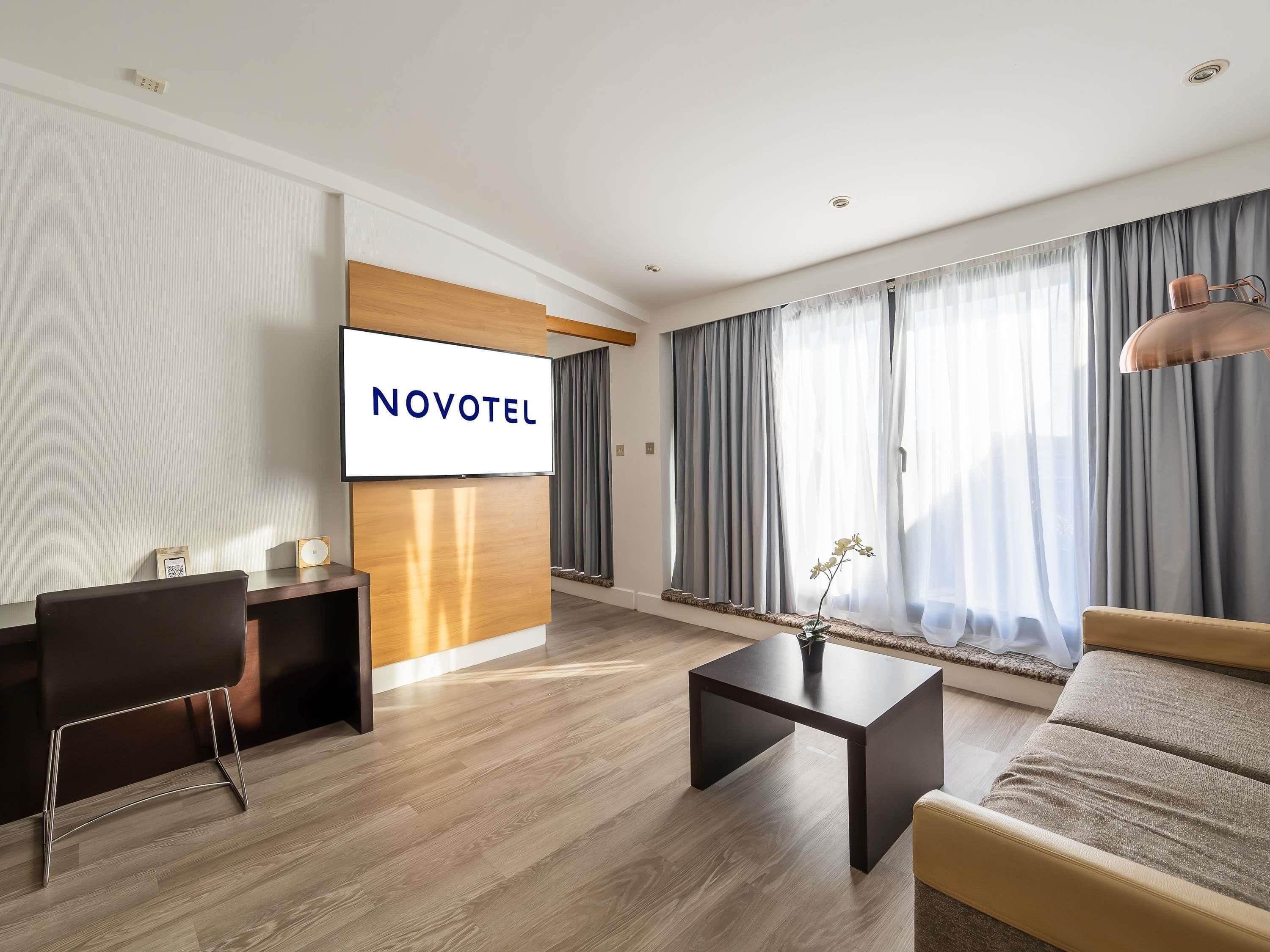 A modern hotel room with a flat-screen TV, a wooden accent wall, a sofa, a coffee table, and large windows with curtains.