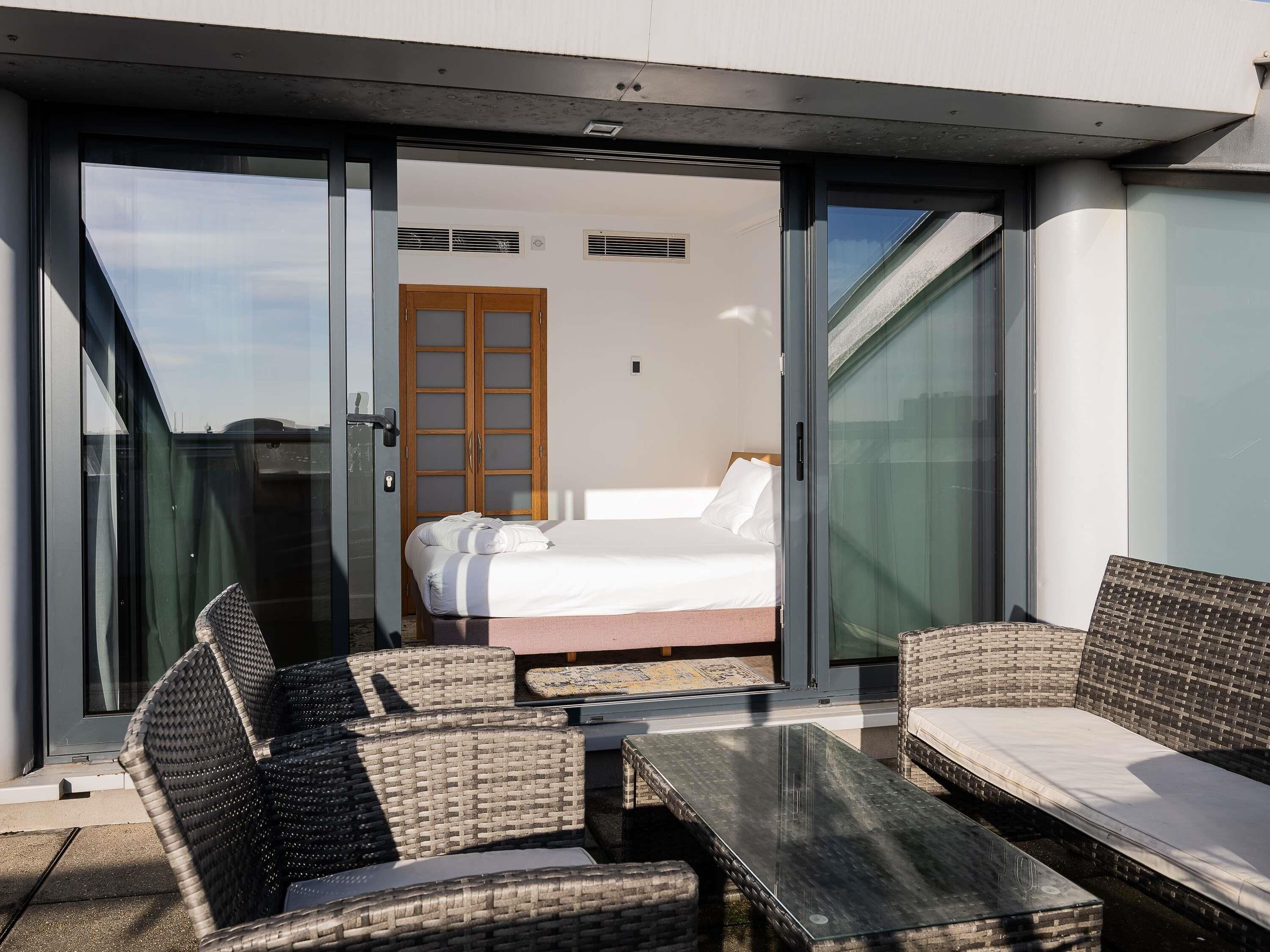 A modern room with a bed, a glass door leading to a balcony, wicker furniture, and a glass table.