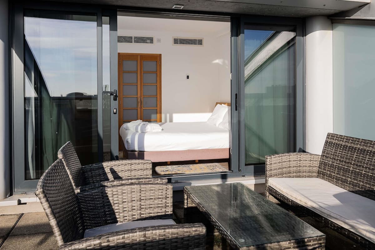 A modern room with a bed, a glass door leading to a balcony, wicker furniture, and a glass table.