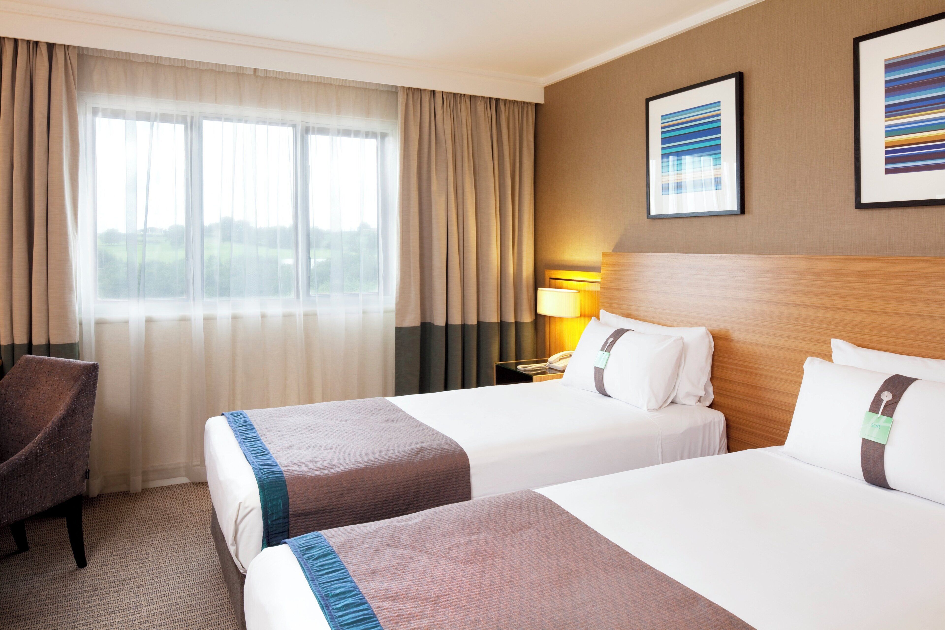 A hotel room with two beds, a chair, a nightstand with a lamp, and a window with curtains.
