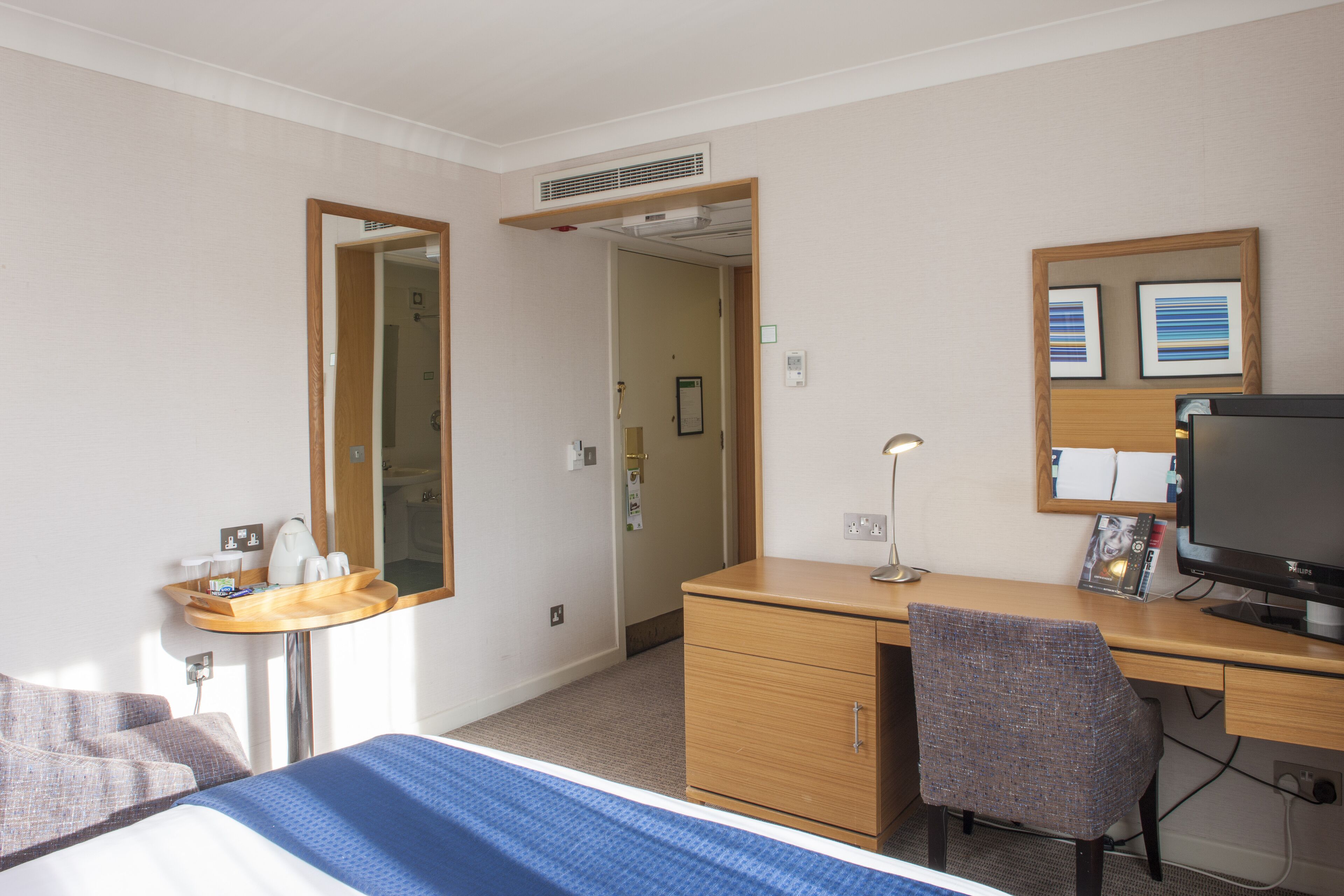 Habitación de hotel con cama, escritorio, silla, espejo, televisión y una mesita con bandeja.