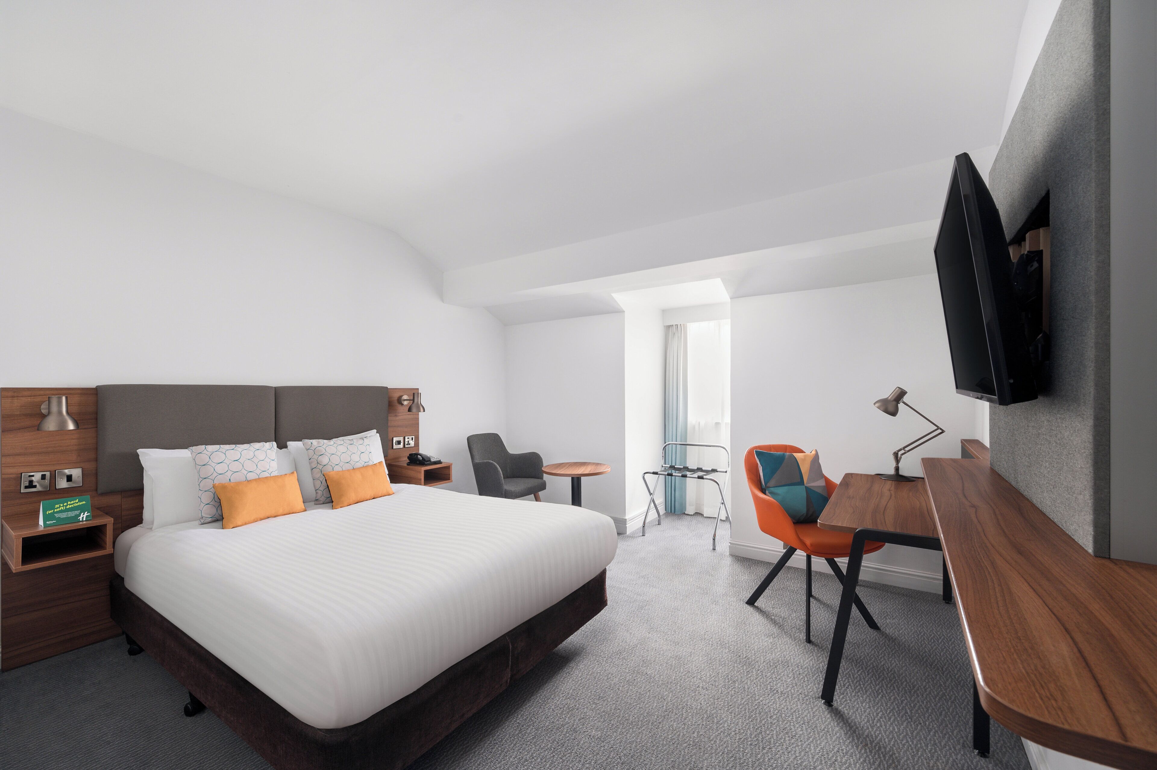 A modern hotel room with a large bed, a desk, a chair, and a TV mounted on the wall.
